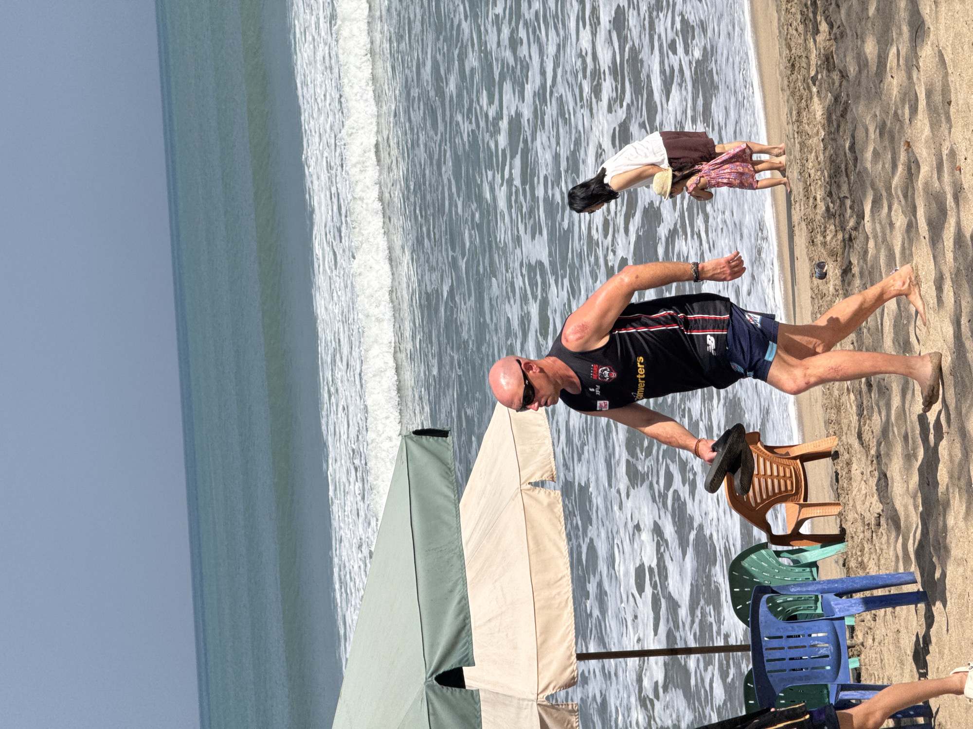A man in a black tank top and blue shorts walks on a sandy beach in Kuta, Indonesia, carrying a pair of flip-flops. He is bald and wearing sunglasses. Behind him, a woman in a white top and brown skirt stands near a young girl in a pink dress and a straw hat. They are at the edge of the ocean, where waves are breaking. Two beach umbrellas, one light beige and one greenish-gray, are visible on the sand, along with several plastic chairs. The sky is clear and blue, and the ocean is a deep turquoise.