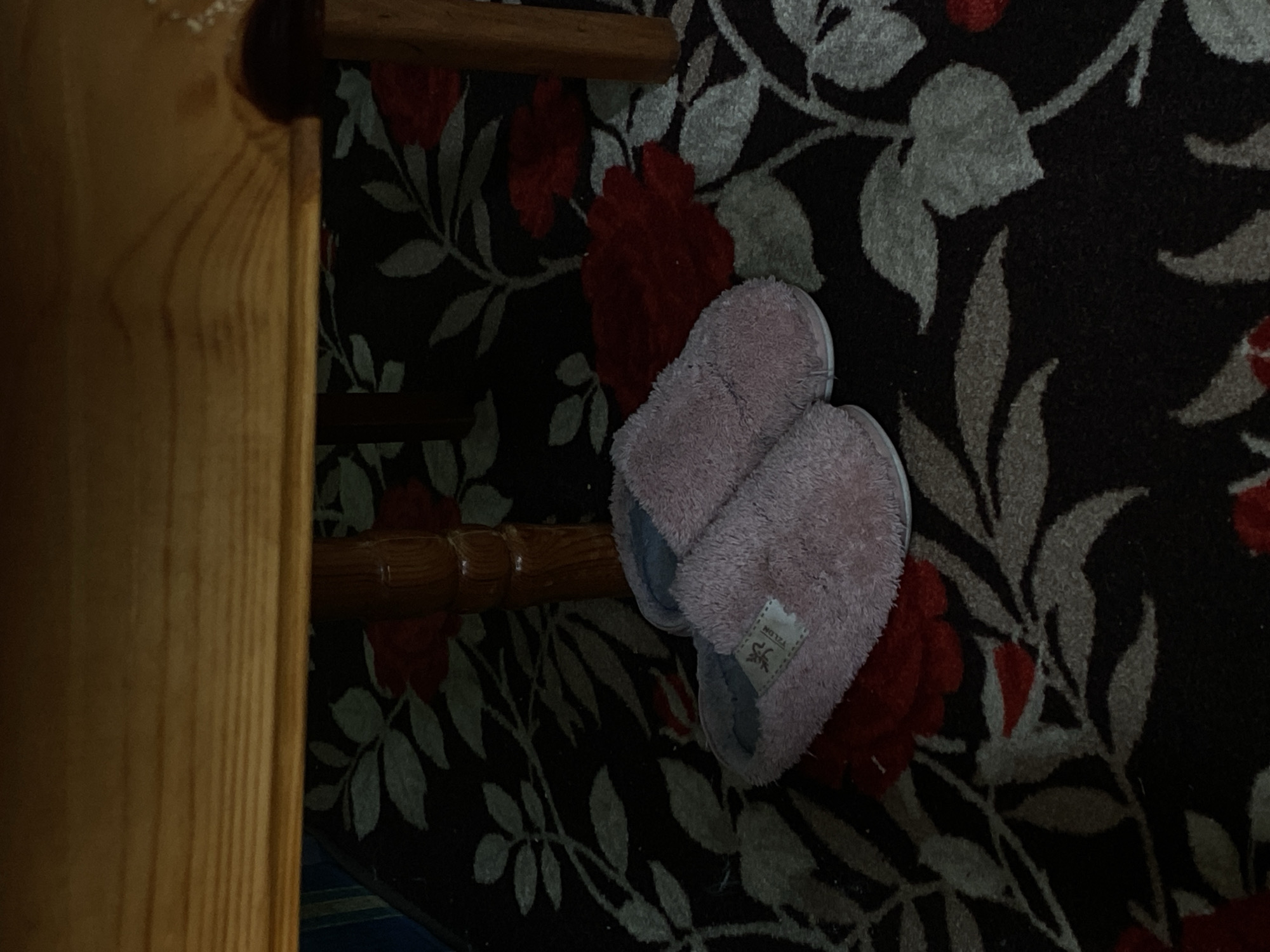 A pair of pink slippers rests on a floral patterned rug next to a wooden stair railing. The rug features a dark background with red and gray floral motifs. The image appears to be taken indoors, near a staircase. There is no information available to determine the location in Blagoevgrad, Bulgaria. No people are visible.