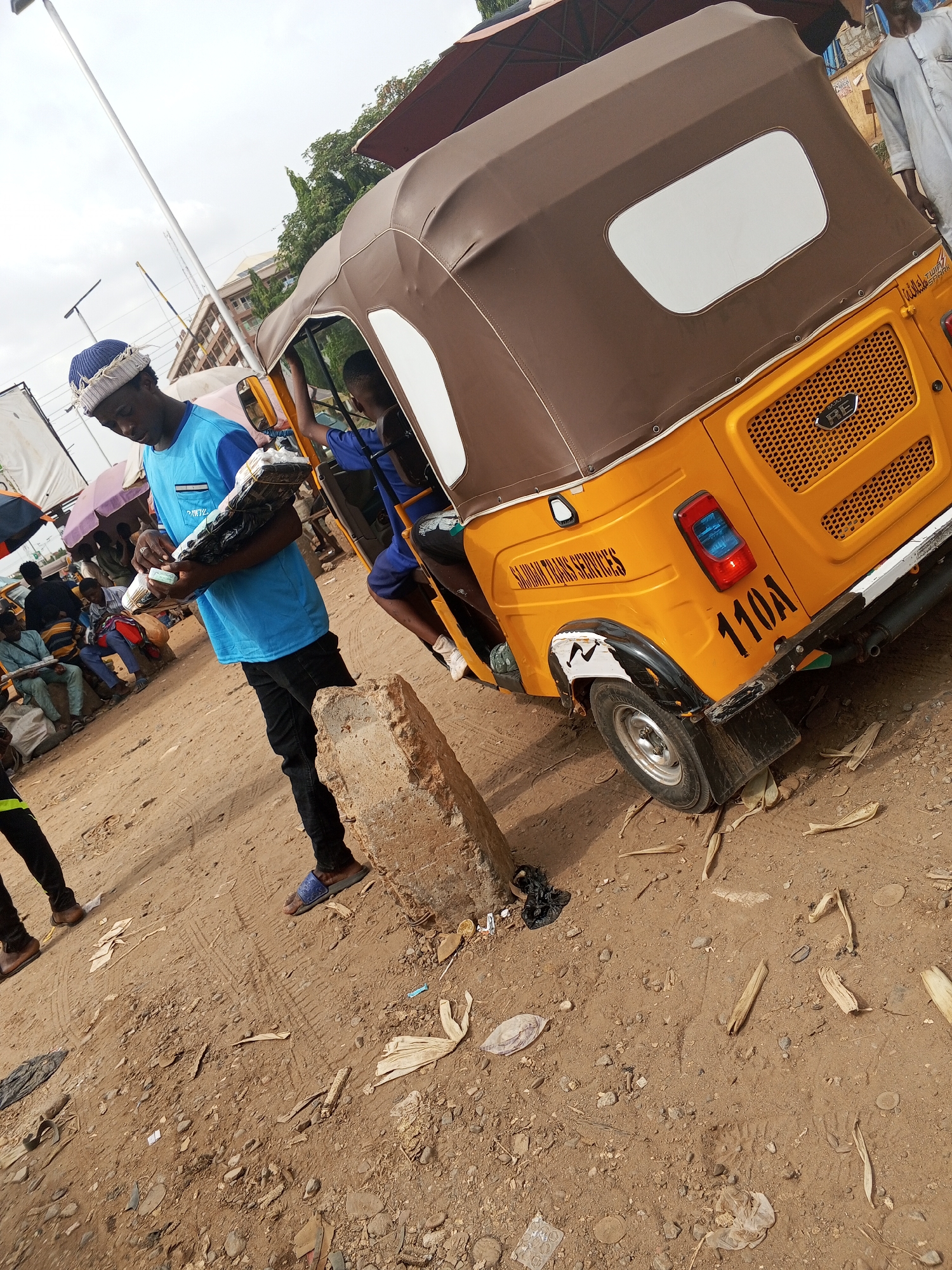 The image depicts a street scene in what appears to be a bustling commercial area of Abuja, Nigeria. A young man wearing a blue t-shirt, black jeans, and a blue and white patterned cap stands on a dirt ground littered with debris. He holds what appears to be a stack of items wrapped in plastic. To his right is a yellow "keke", a three-wheeled motorized vehicle common in Nigeria, with a brown canvas top. The keke bears the identification number "110A" and the phrase "SANMAN TRANS SERVICES". Inside the keke, another person is visible. In the background, other people are sitting and standing under umbrellas. The ground is primarily dirt and gravel.