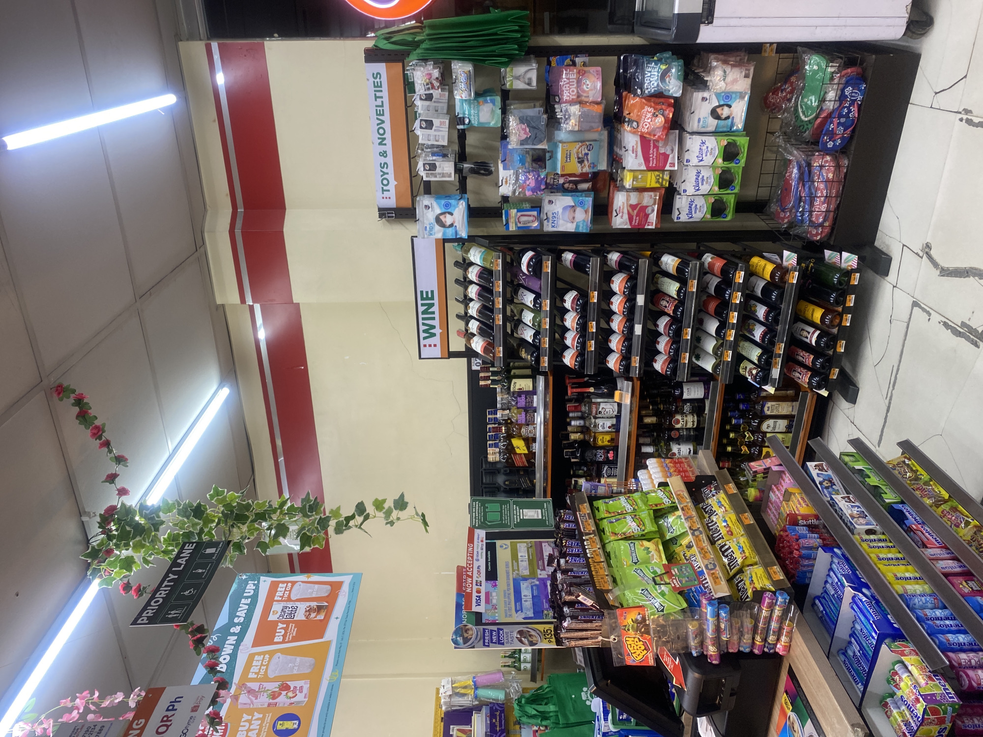 This image shows the interior of a convenience store, likely a 7-Eleven branch, in Davao City, Philippines. The scene is brightly lit by fluorescent lights and features shelves stocked with various products, including alcoholic beverages, snacks, and travel-related items. A prominent "PRIORITY LANE" sign is visible, along with decorative vines and flowers hanging from the ceiling. The store appears to be well-organized and stocked, with no customers visible in the frame. The overall impression is that of a typical convenience store operating in an urban environment.
