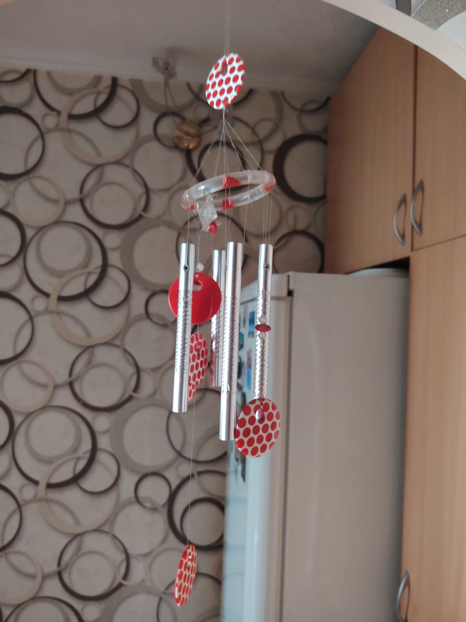 A wind chime with silver tubes and red, polka-dotted decorative elements hangs in front of a patterned wallpaper. To the right of the wind chime, a portion of a white refrigerator and wooden cabinets are visible. The scene is indoors, suggesting a domestic setting. The image was taken in Rostov-on-Don, Russia.