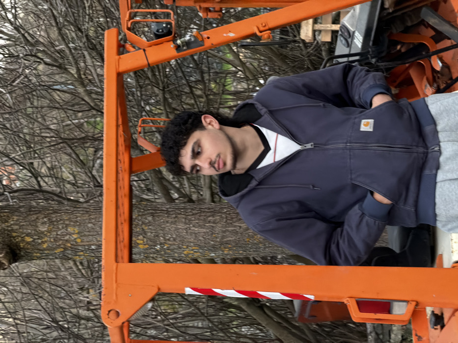 The image displays a young adult male standing outdoors in the city of Bragança, Portugal. The individual has dark, curly hair, a mustache, and a short beard. He is wearing a navy blue zippered hooded jacket with a visible Carhartt logo on the left pocket, over a white v-neck shirt with black trim and a red stripe, and gray sweatpants. His hands are inserted into the pockets of his jacket. His gaze is directed towards the right side of the frame. The subject is positioned in front of a prominent bright orange metal structure, likely part of heavy machinery or scaffolding. A vertical support beam of this structure, featuring a red and white diagonal striped reflector, is visible on the left. More orange structural elements and a black component are visible to the right of the individual. The background consists of numerous bare deciduous tree branches and a tree trunk displaying textured bark and lichen patches.