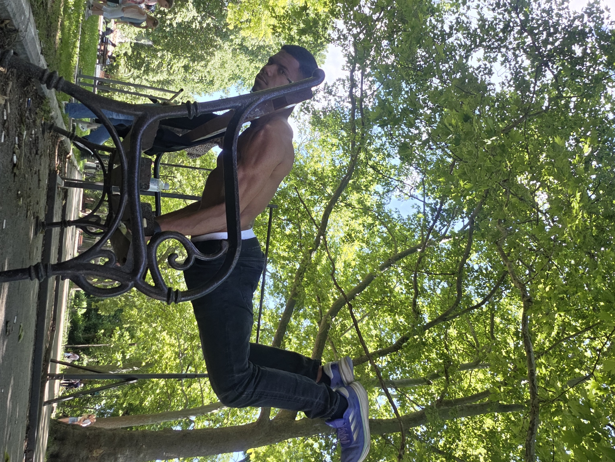 A young adult male, appearing to be in his late teens or early twenties, is performing a difficult calisthenics exercise. He is shirtless, revealing a muscular physique, and wearing dark jeans and athletic shoes. His body is extended horizontally, with his hands on the back of a dark, ornate metal park bench, and his legs are lifted and bent at the knees, resting on the back of the bench as well, or possibly on another unseen surface. The exercise appears to be a variation of a planche or a similar maneuver. He is positioned outdoors in a park setting, under the dappled shade of large, leafy trees, suggesting a warm, sunny day. The background shows a pathway, other park benches, and a few other individuals in the distance, including two young girls and some adults. The scene is likely situated in Varna, Bulgaria, given the context provided. The overall impression is one of athleticism and strength displayed in a natural, public space.