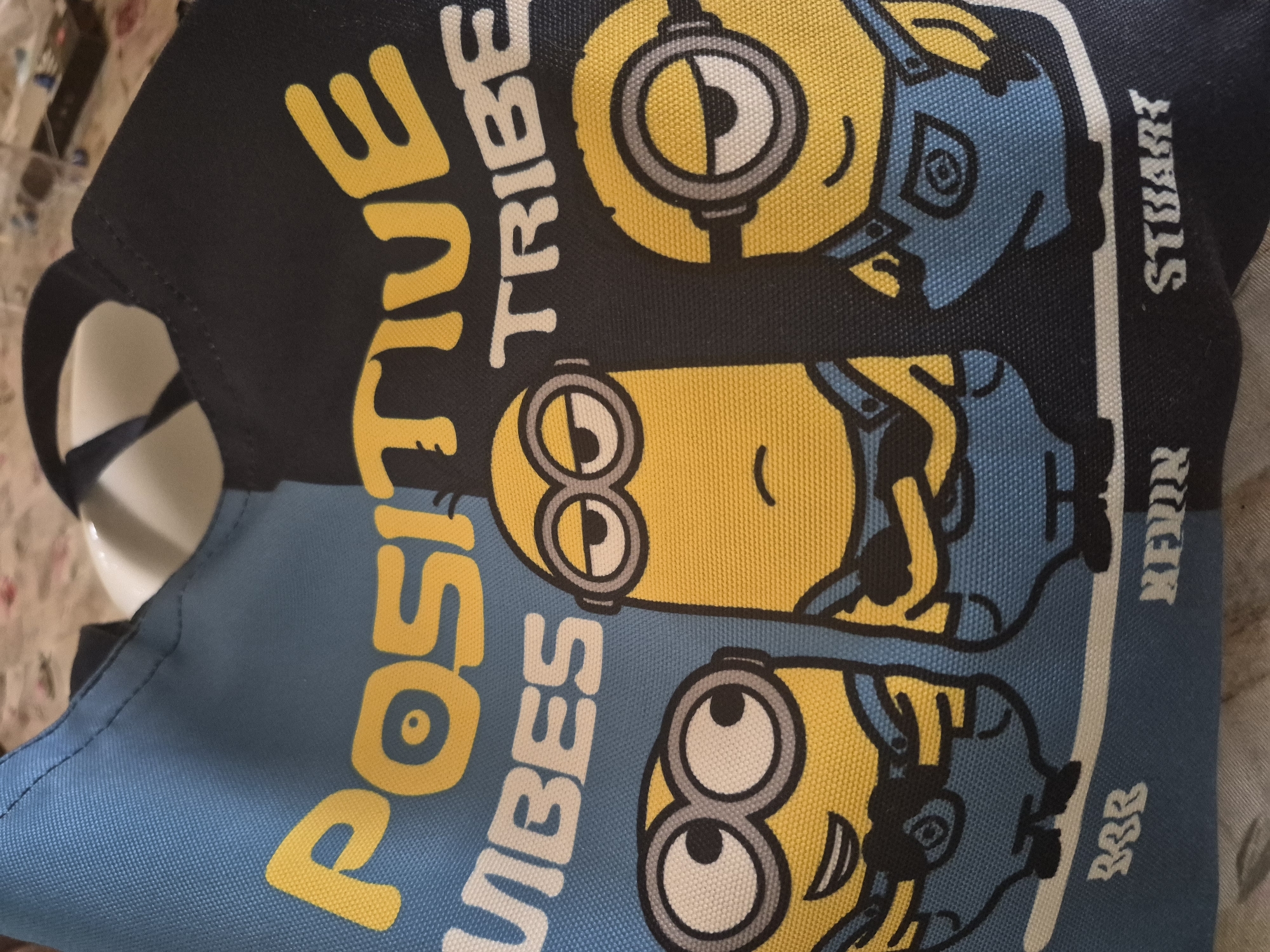 This media file displays a close-up, angled view of a fabric bag, likely a tote or reusable bag, featuring a vibrant design. The bag is divided horizontally with a dark blue upper section and a lighter blue lower section.
Prominently featured are three yellow Minions characters from the Despicable Me franchise, each with black outlines and white and gray goggles. From top to bottom, the Minions are identified by their names printed vertically in white text on the right side of the bag: "STUART" (a one-eyed Minion), "KEVIN" (a two-eyed Minion looking slightly upwards), and "BOB" (a two-eyed Minion with a grinning expression).
On the left side of the bag, bold yellow text reads "POSITIVE" vertically, with white text "TRIBE" beside it. Below "POSITIVE", "VIBES" is printed in white, also vertically. The fabric has a visible weave texture. The background is blurred, suggesting an indoor setting.