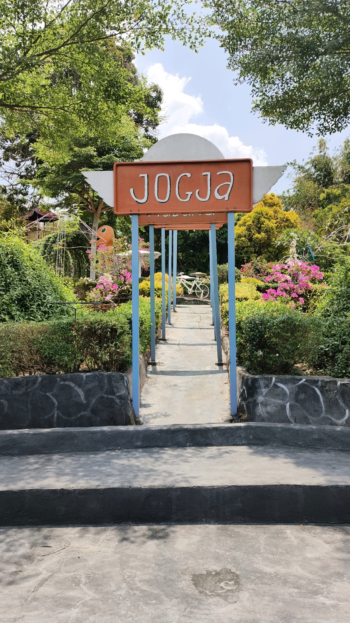 In Air Paoh, Indonesia, a path leads through a vibrant garden. A prominent sign with the word "JOGJA" in white letters against an orange background, framed by white wings, marks the entrance. The path, paved with light grey concrete, is lined on both sides by lush green bushes and colorful flowers, predominantly pink and yellow. Blue posts support a canopy over the path, leading towards a white bicycle parked in the background. The scene is set on a clear day with blue skies and scattered clouds, with trees and foliage surrounding the garden. The ground in the foreground consists of concrete steps and a textured concrete area.