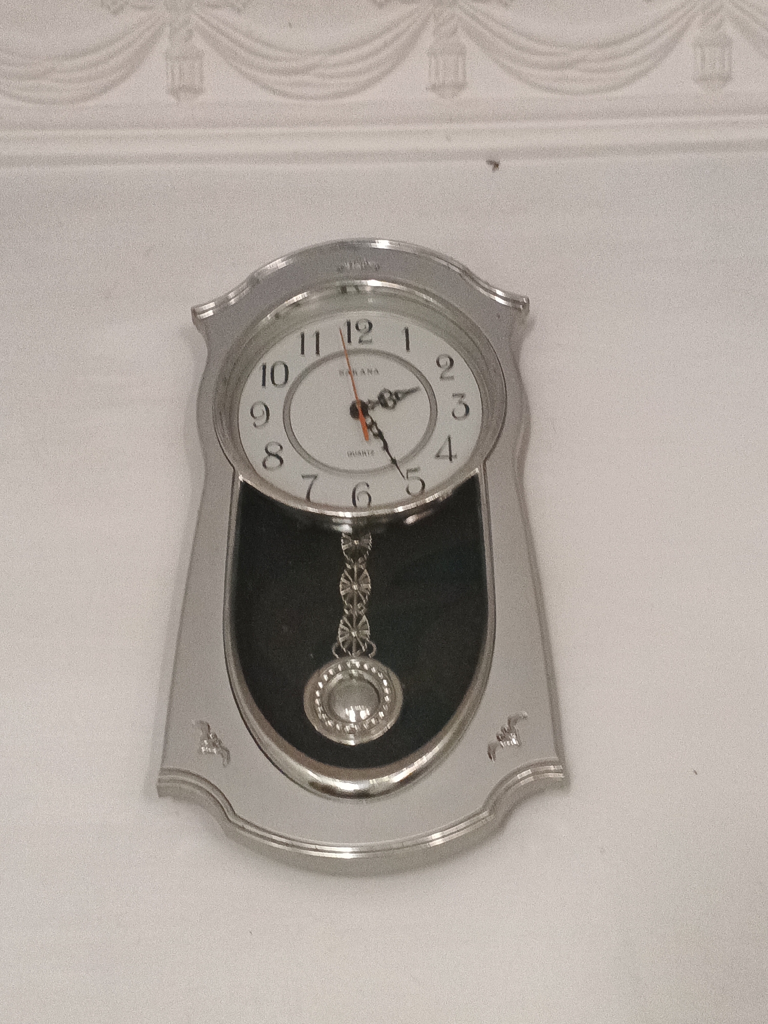 A silver pendulum clock with a white face is mounted on a light-colored wall. The clock has black Arabic numerals for the hours, with the numbers 1 through 12 clearly visible. The hour hand is pointing slightly past the 5, and the minute hand is pointing towards the 12. A decorative silver chain hangs from the bottom of the clock, holding a silver pendulum at the end. The wall appears to have some decorative molding or detailing at the top. The image is taken from a straight-on perspective, slightly below the clock. The overall lighting is even, with no harsh shadows.
