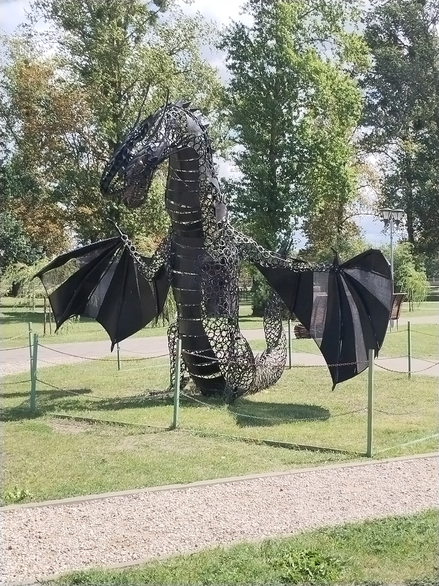 An outdoor sculpture of a black dragon made of metal chains and rings stands in a grassy park area. The dragon is in a dynamic pose with its wings spread wide. The park has trees in the background, a gravel path in the foreground, and decorative chains and posts surrounding the sculpture. The sky is partly cloudy. This sculpture is located in Brest, Belarus.