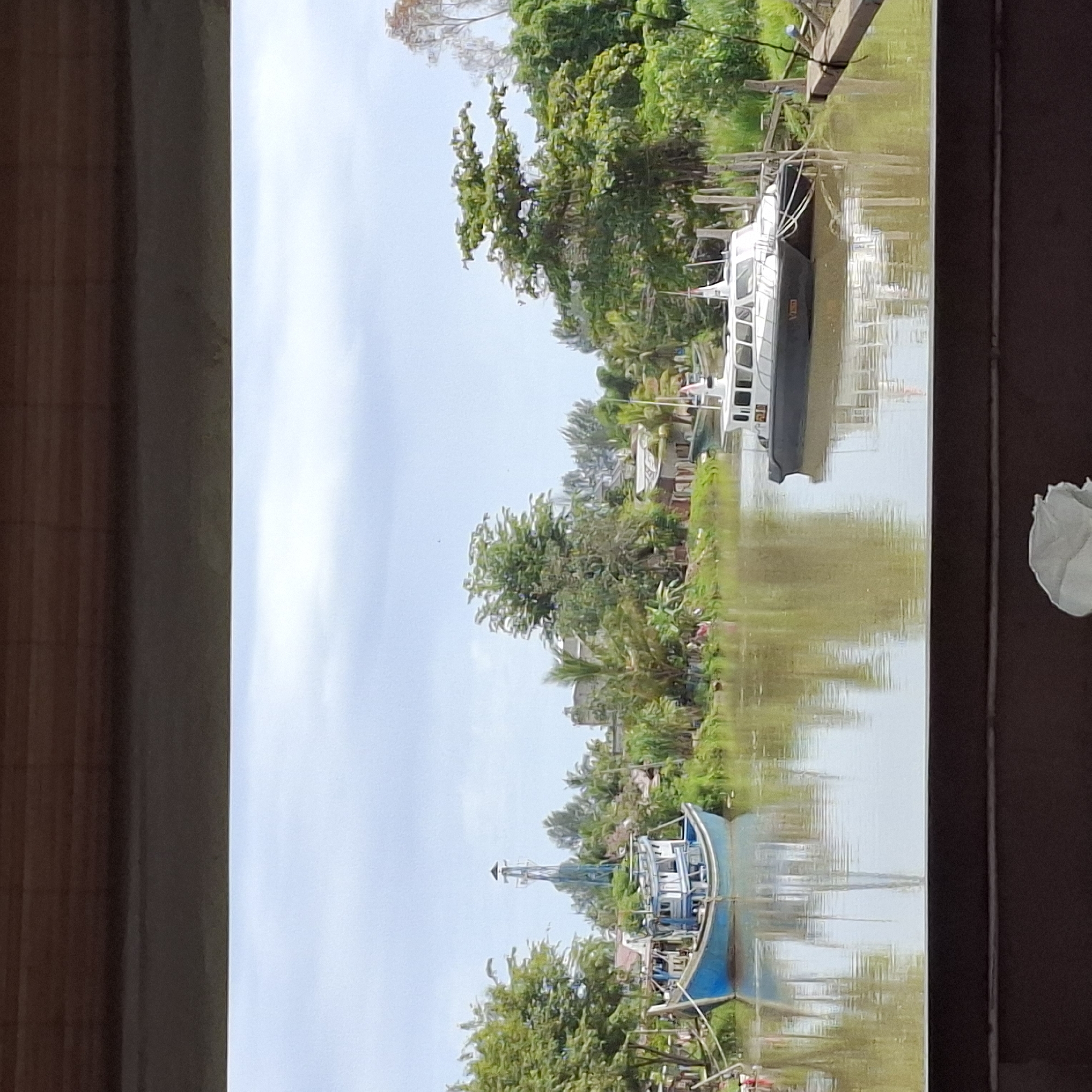 The image captures a serene canal scene in Mesjid Tuha, Indonesia. The canal, with its murky brown water, reflects the overcast sky and the lush green vegetation lining its banks. Two boats are the primary subjects. A blue and white fishing boat is moored to the left, its weathered exterior suggesting it is actively used. To the right, a sleek white and dark-hulled patrol boat is anchored, its presence hinting at security or enforcement operations. The background reveals a cluster of buildings, some appearing to be houses, interspersed with abundant tropical trees and palm fronds, characteristic of a coastal or riverside settlement. A tall, slender structure, possibly a lighthouse or a communication tower, stands behind the fishing boat, adding to the unique landscape. The foreground is framed by dark, horizontal wooden elements, suggesting the photo was taken from a covered vantage point or a structure overlooking the canal. No people are clearly visible in the image, contributing to the tranquil atmosphere.