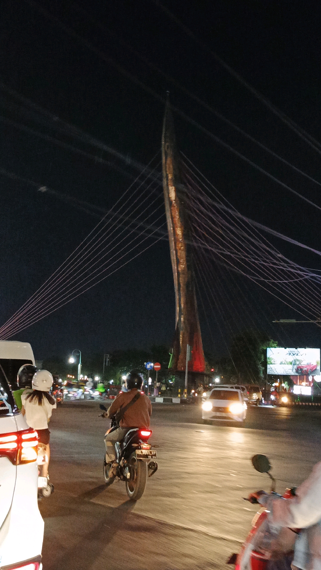 The image captures a nighttime scene at the Semanggi bridge in Surakarta, Indonesia. A man wearing a brown shirt and helmet is riding a motorcycle in the foreground, his license plate visible. Two people on a motorcycle, one with a white helmet, are positioned to the left, facing away from the camera. Several vehicles are visible on the road, with headlights illuminating the scene. In the background, the distinctive structure of the Semanggi bridge stands tall, adorned with numerous cables that extend upwards. Streetlights and an illuminated billboard are also present. The overall atmosphere is that of a busy urban environment at night.