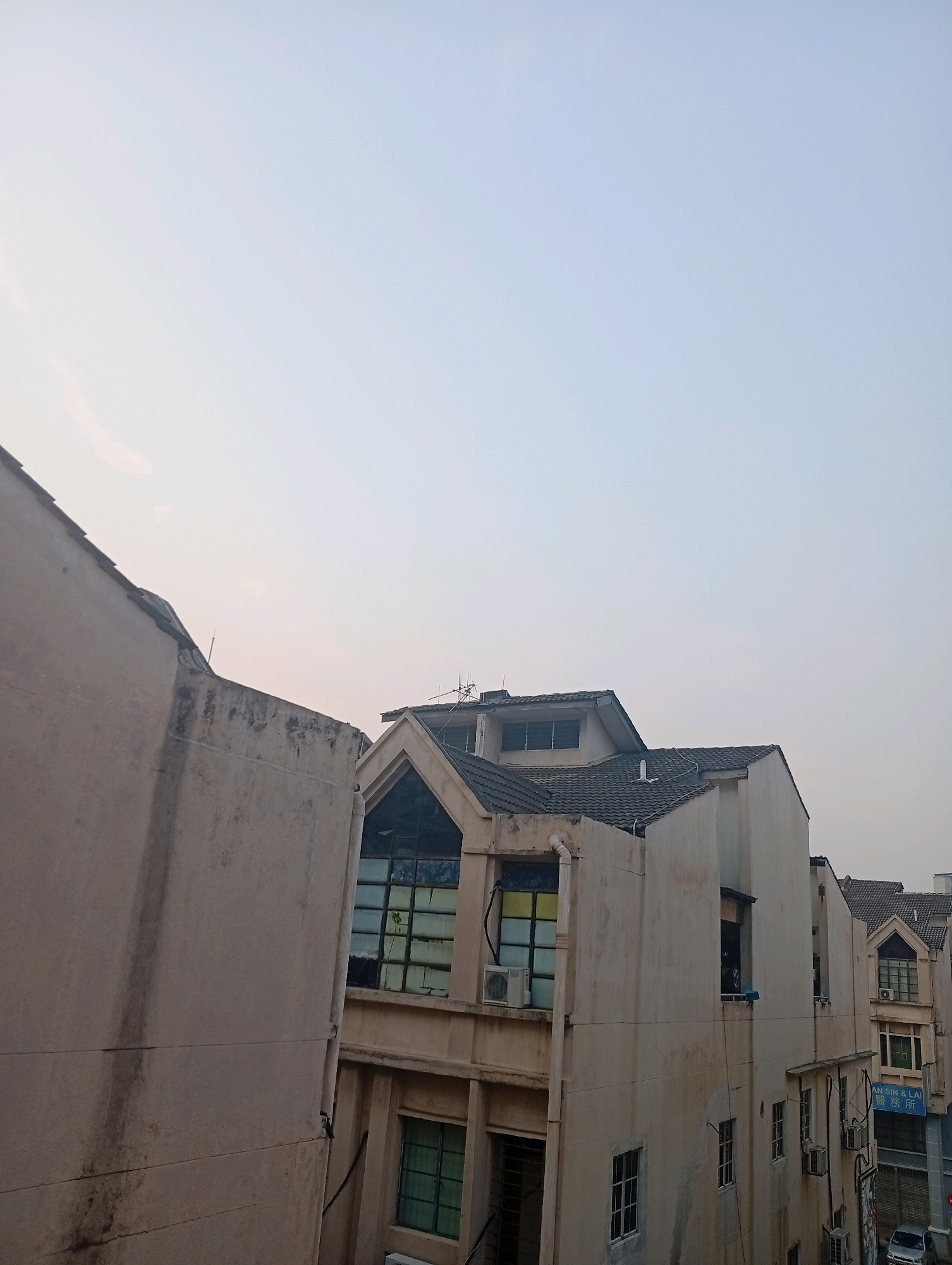 In Selayang Municipal Council, Malaysia, the image captures a row of multi-story buildings under a pale, overcast sky. The buildings, primarily cream-colored, exhibit signs of age with visible discoloration and some peeling paint. Their architectural styles vary, with some featuring pitched tile roofs and distinctive gabled windows, while others have flatter, more utilitarian designs.
Several windows are visible, some with closed blinds or panes, others showing glimpses of interior spaces. Air conditioning units are mounted on some of the building facades. A blue sign with Chinese characters is affixed to one of the buildings, suggesting a commercial establishment. Power lines and conduits run along the exteriors.
In the foreground, to the left, is a portion of a building with a textured, light-colored wall. The overall impression is of an urban street scene, possibly a residential or mixed-use area, with a slightly worn but functional appearance. The lighting suggests it might be early morning or late afternoon due to the soft, diffused sunlight.