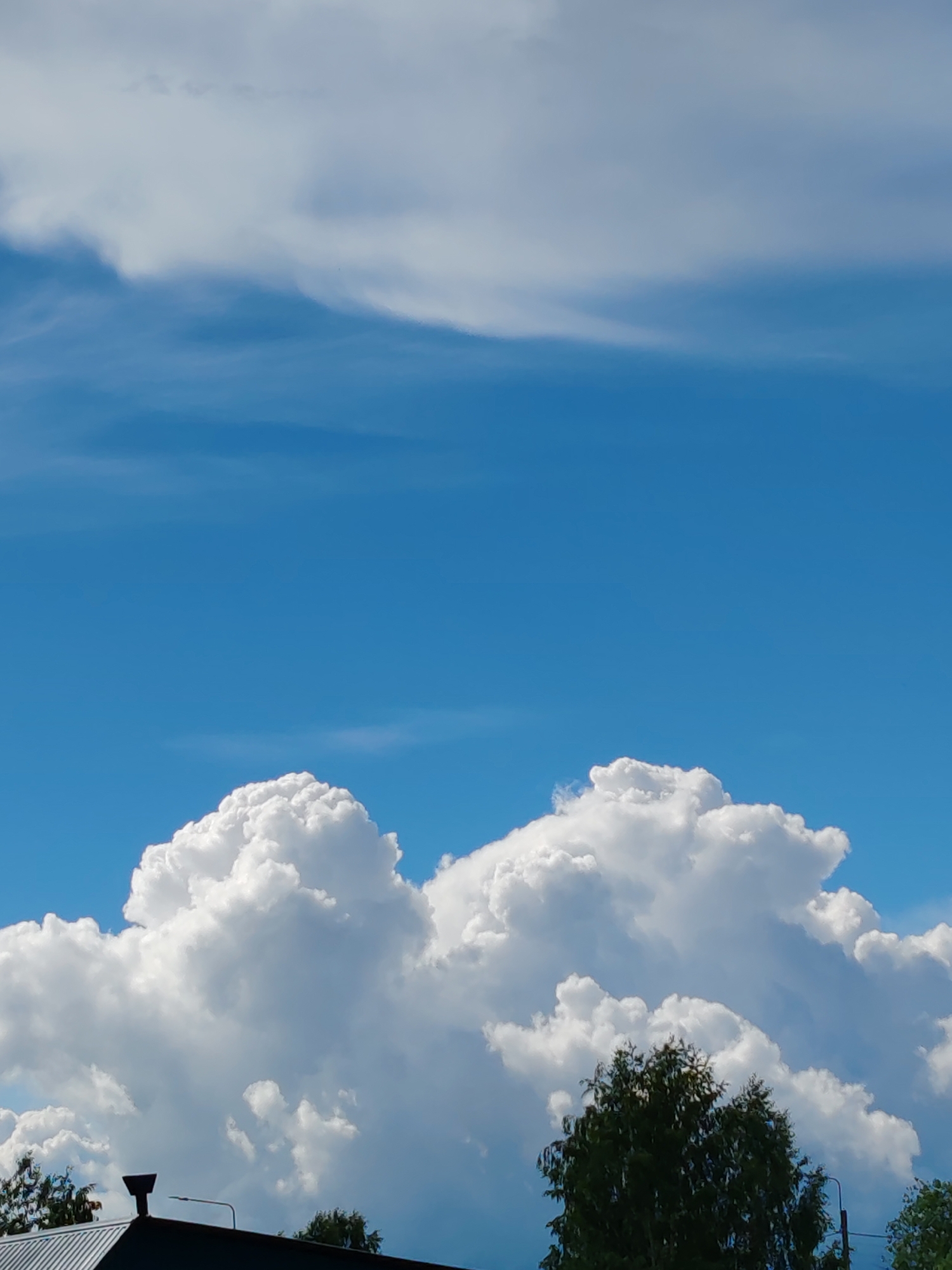 The image depicts a clear blue sky with scattered white cumulus clouds, typical of a sunny day. The bottom portion of the image shows the rooftops of several buildings and the tops of trees. One rooftop is prominently visible in the foreground with a dark-colored, angled surface, possibly a shingled roof. A black chimney-like structure is situated on this roof. A streetlamp is also visible extending from the roofline. In the mid-ground, the lush green foliage of trees extends upwards, obscuring parts of the sky. The lighting suggests it is daytime, with the sun illuminating the clouds and foliage. The scene appears to be an outdoor view, likely from a residential or urban area.