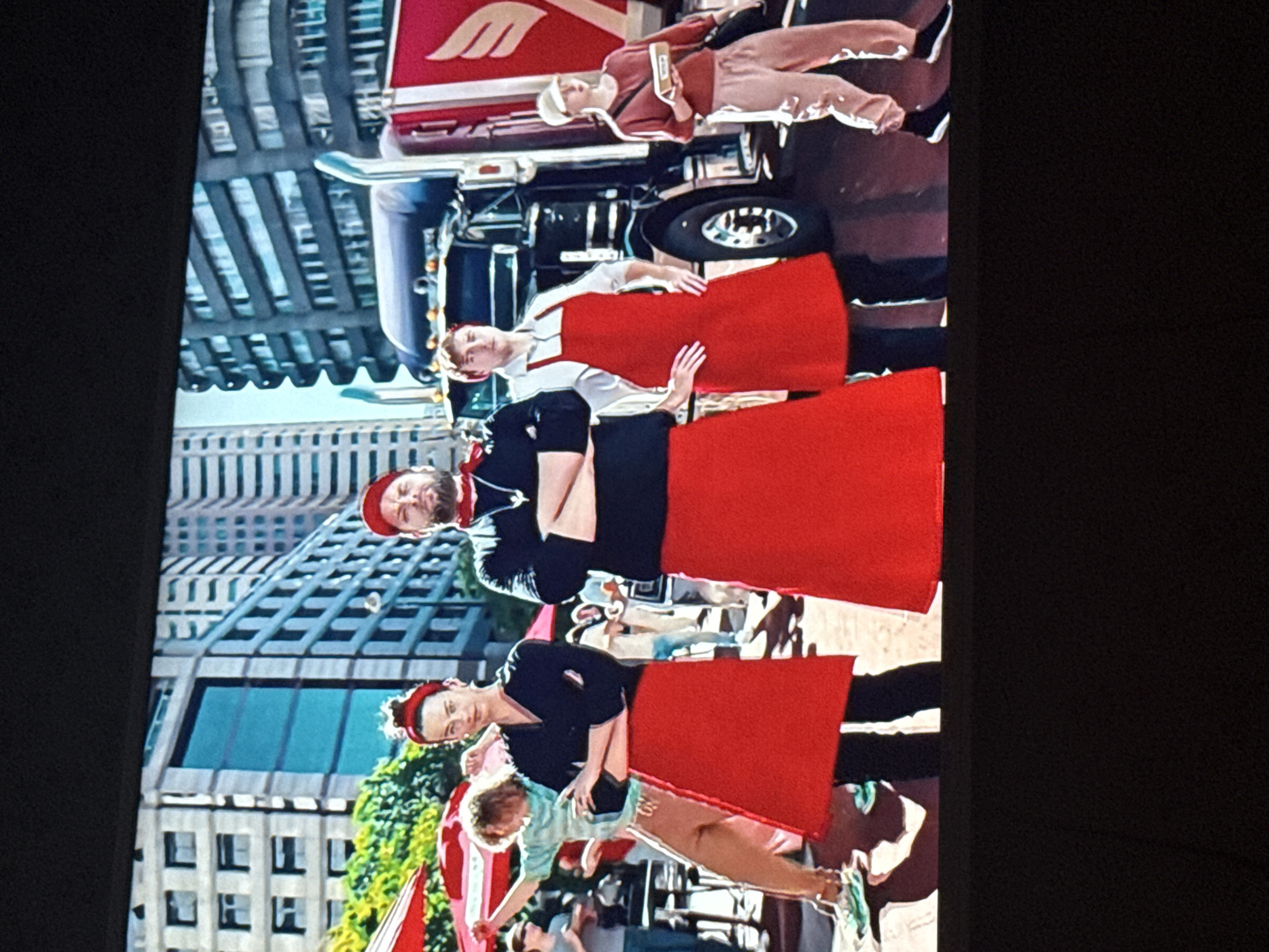 The image depicts a scene with several people standing in front of tall buildings, possibly in a city. Three people in the foreground wear dark tops and red aprons. There's a younger person wearing a light blue top. One person is on top of a truck. Tall buildings are visible in the background. The image lacks the details to confirm the specific location in Mytishchi, Russia.