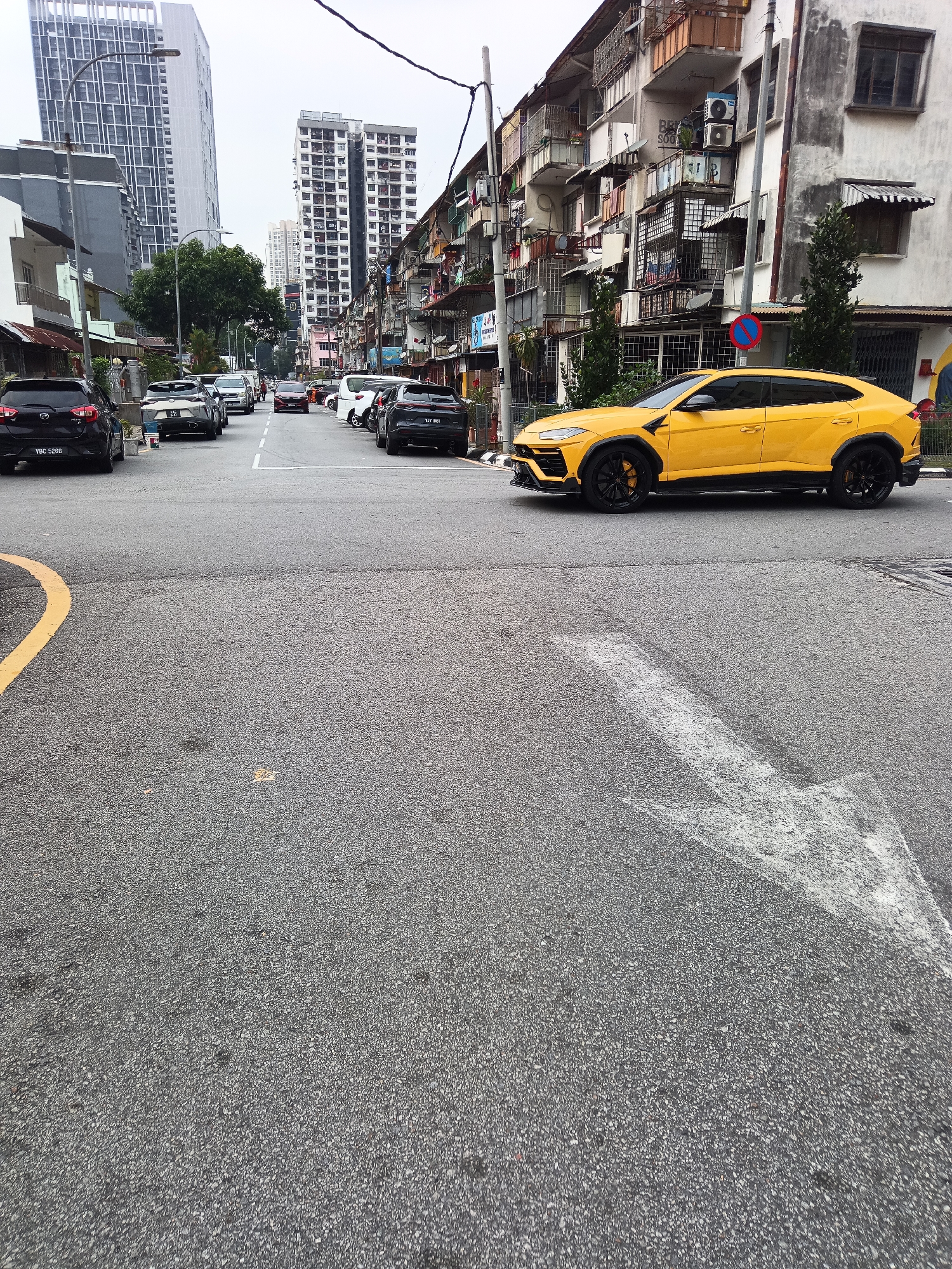 A bright yellow Lamborghini Urus is parked on an asphalt road, its front bumper extending over a white directional arrow painted on the ground. The car is positioned in the foreground, slightly to the right of the frame, with its driver-side door facing the viewer. The vehicle is sleek and modern, with black accents on its wheels, side skirts, and front splitter. Behind the car, the road continues into the distance, lined with parked vehicles and buildings. To the left of the road, a black Perodua Myvi with license plate VBC 5266 is visible. Further down the road, several other cars are parked, including a white van and a silver Lexus.
The buildings on the right side of the street are old, multi-story apartments with balconies and weathered facades. The building in the far right has an air conditioning unit mounted on its exterior wall. A blue and red circular traffic sign with a diagonal red line is affixed to a pole in front of the apartment building. To the left of the road, modern high-rise buildings dominate the skyline, including a tall, slender skyscraper with a distinct architectural design. The sky is overcast, contributing to a muted light across the scene. The overall environment suggests an urban setting within Kuala Lumpur, Malaysia.