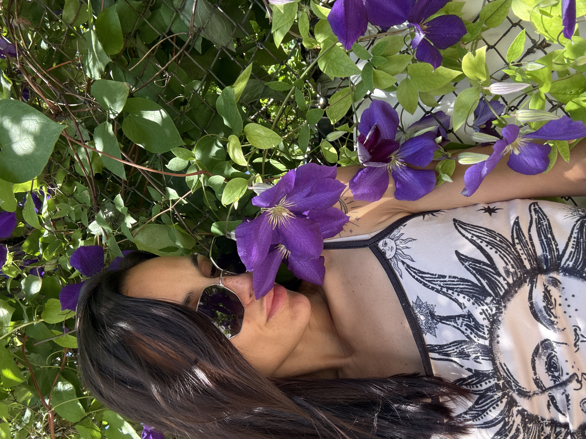 This is an outdoor, eye-level, medium shot of a woman resting amongst purple clematis flowers and green foliage, which are climbing on a chain-link fence. The woman, with dark hair and wearing sunglasses and a white top with a black sun graphic, is partially obscured by the flowers and her hair. Her face is turned towards the camera, with her eyes closed. The sun is shining brightly, casting shadows on the leaves and illuminating the scene, suggesting it is daytime. The overall mood is peaceful and serene. The location is identified as Pleven, Bulgaria.