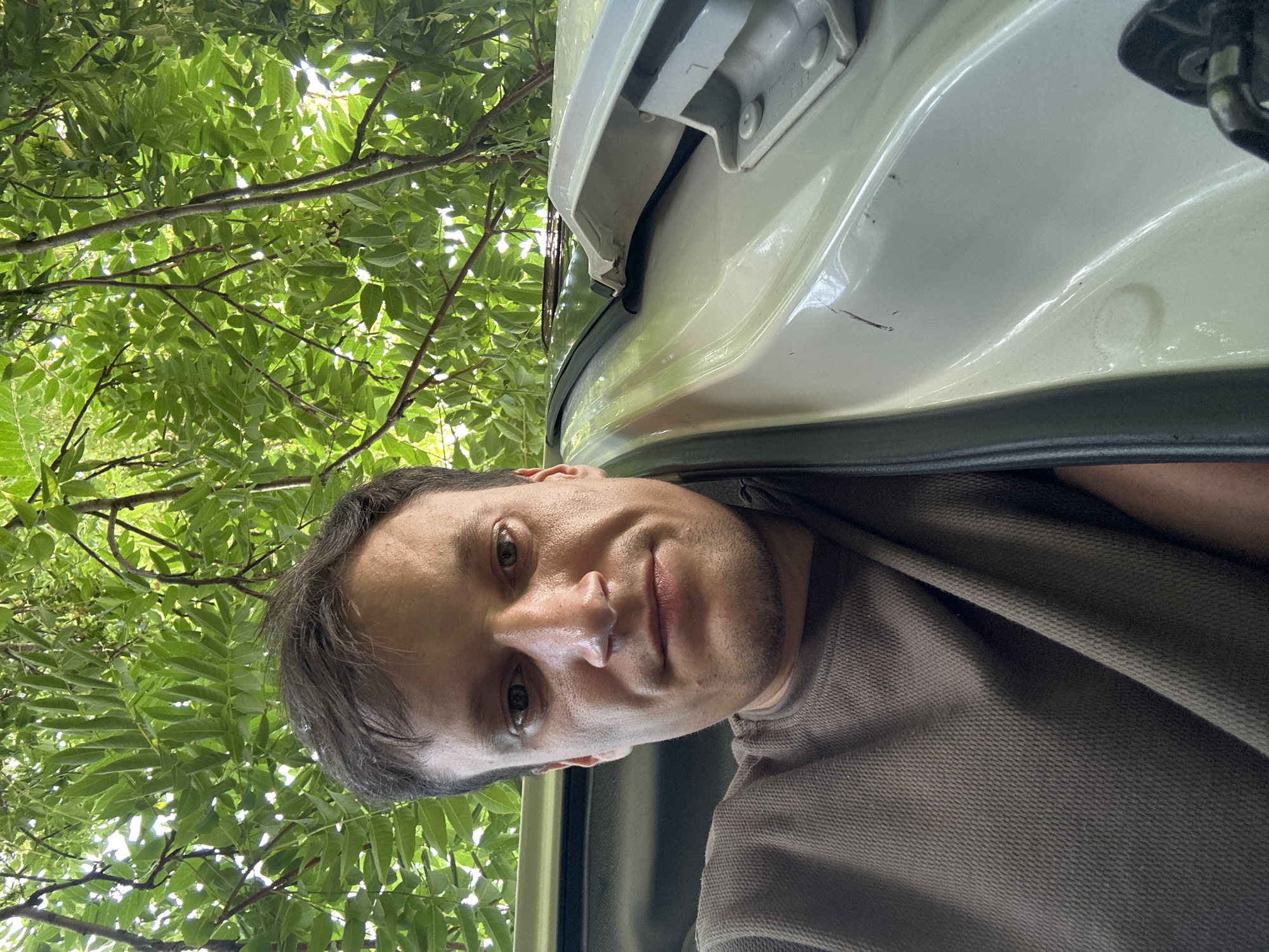 The image is a low-angle, close-up selfie of a man with short, dark hair and stubble, wearing a brown, textured t-shirt. He is positioned in the doorway of a white vehicle, with the car's interior visible on the right side of the frame. Overhanging branches and green foliage of a tree dominate the background, suggesting an outdoor setting. The overall lighting indicates daylight. The specific location in Makiv, Ukraine, is not determinable from the visual information provided.