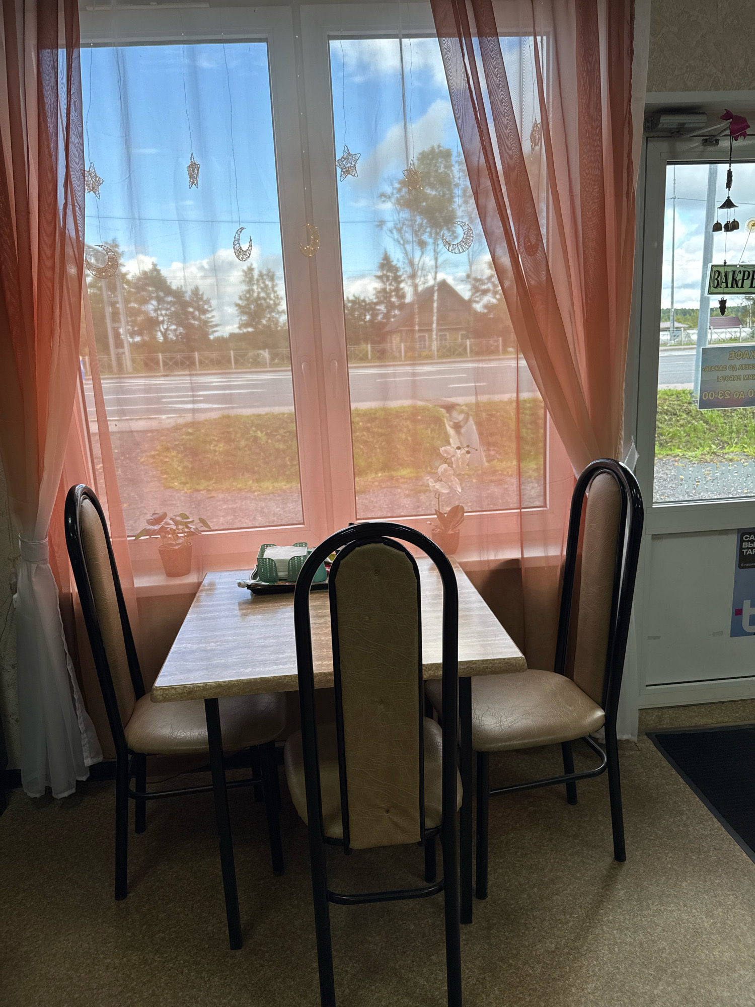 An interior shot shows a small dining area with a square table and three chairs. The table is light-colored with a speckled surface and is set with a tray of napkins and a small potted plant on the windowsill. Two chairs are positioned on the left side of the table, and one is on the right. A third chair is positioned behind the table, facing away from the camera. The chairs have metal frames and light brown upholstery.
Behind the table, a large window offers a view of a road and trees. The window is adorned with sheer, peach-colored curtains that are pulled to the sides, revealing string lights with star and crescent moon shapes. The exterior scene appears to be a rural or suburban area with a road, grassy areas, and trees.
To the right of the window, a glass door leads to an exterior area. A sign hanging on the door, partially visible, reads "ЗАКРЫТО" (CLOSED) in Russian. Another sign below it has Cyrillic text. The floor inside is carpeted with a flecked pattern. The overall impression is of a small, simple dining space, possibly in a cafe or restaurant in Ryabovo, Russia, that is currently closed.