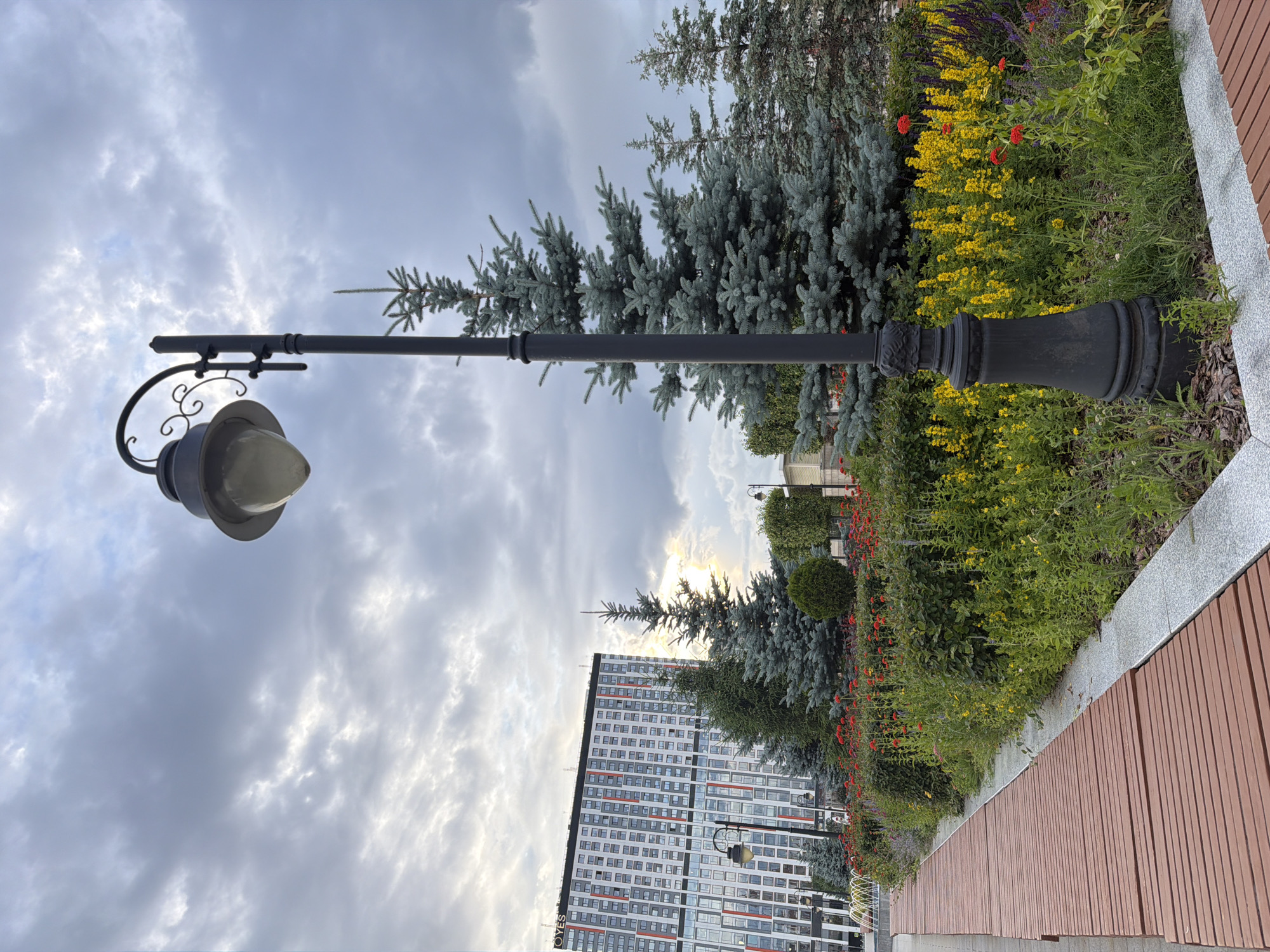 A wide shot captures an outdoor public space with a wooden walkway in the foreground on the left, leading towards a building. A black lamppost stands to the right of the walkway, featuring an ornate base and a glass lantern. Behind the lamppost is a bed of vibrant flowers, predominantly yellow and red, with lush green foliage and several fir trees. In the background, a tall, modern building with a white facade and red and gray accents dominates the skyline. The sky above is overcast with dramatic, textured clouds. The scene appears to be a park or urban plaza, possibly in a residential or commercial area given the building's architecture. There are no people visible in the image.