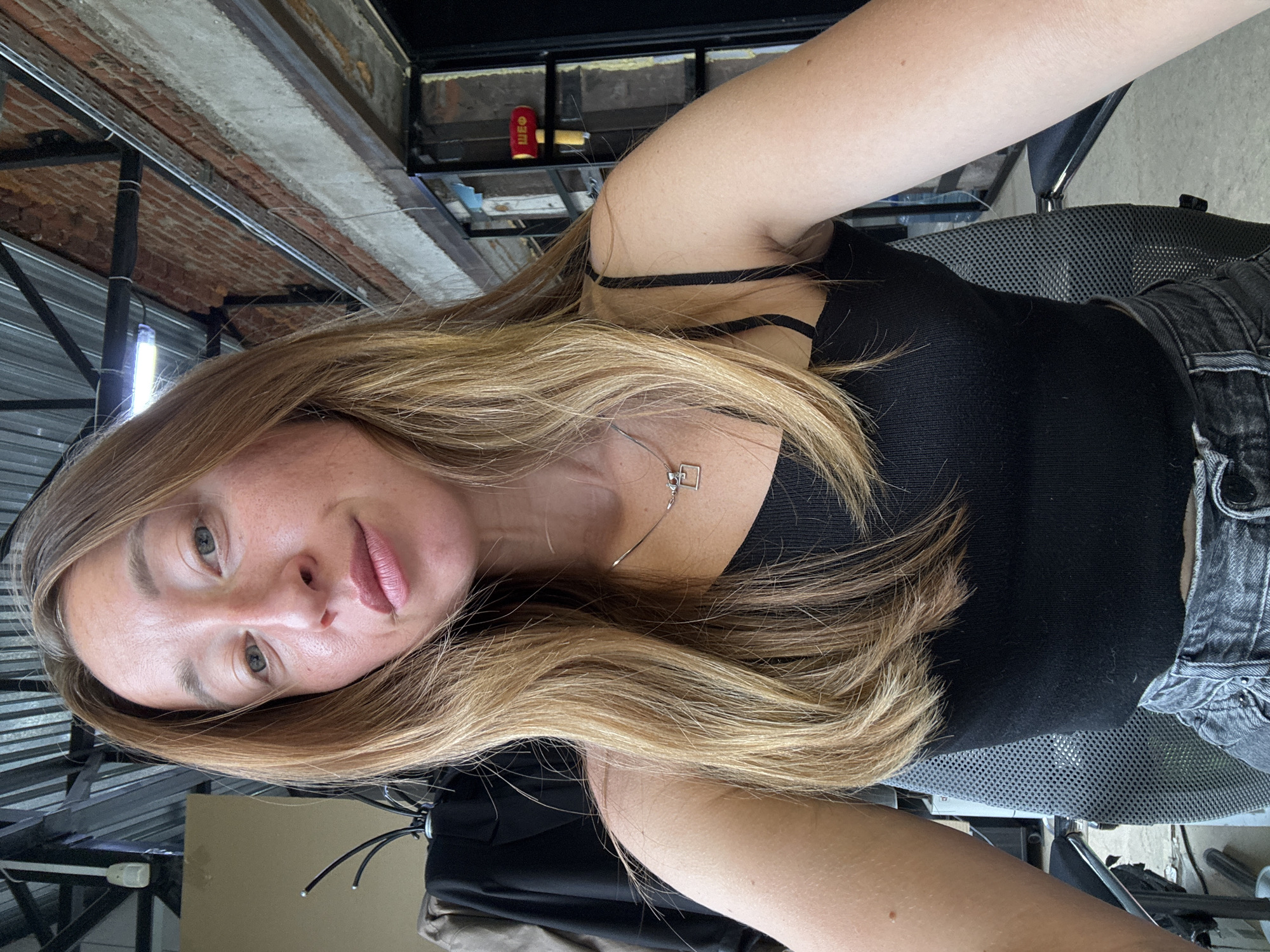A woman with light brown hair and green eyes is centered in the image. She is wearing a black tank top and a silver necklace. She is seated in a grey mesh chair. The background shows a structure with exposed brick, beams, and metal sheeting. A hammer with a red handle and the letters "ШЕО" in yellow is visible on a shelf in the background. This appears to be an indoor setting with industrial characteristics. There is no evidence to confirm the location as Chaikovsky, Russia.