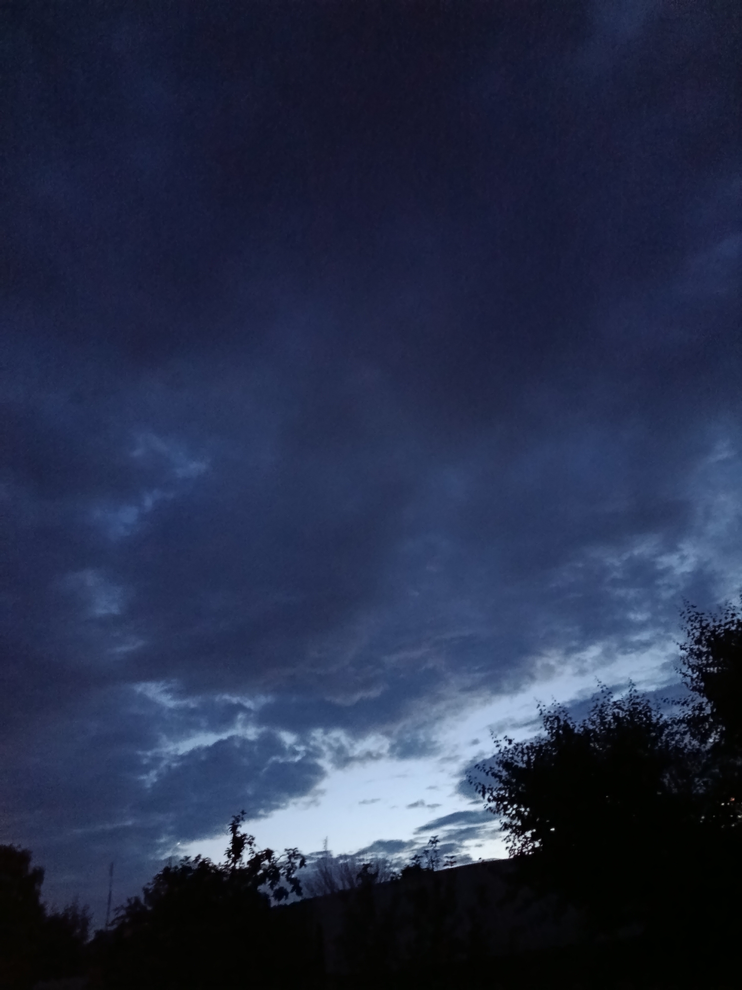 The image depicts a dark, cloudy sky at dusk over the city of Marx, Russia. The sky is predominantly a deep navy blue, with darker clouds obscuring most of the light. Towards the bottom of the frame, there are lighter patches of sky suggesting the horizon. Silhouetted against the sky are trees and what appear to be buildings or structures. The overall impression is one of twilight or an approaching night, with a somber and moody atmosphere. There are no people or identifiable actions visible in the scene.