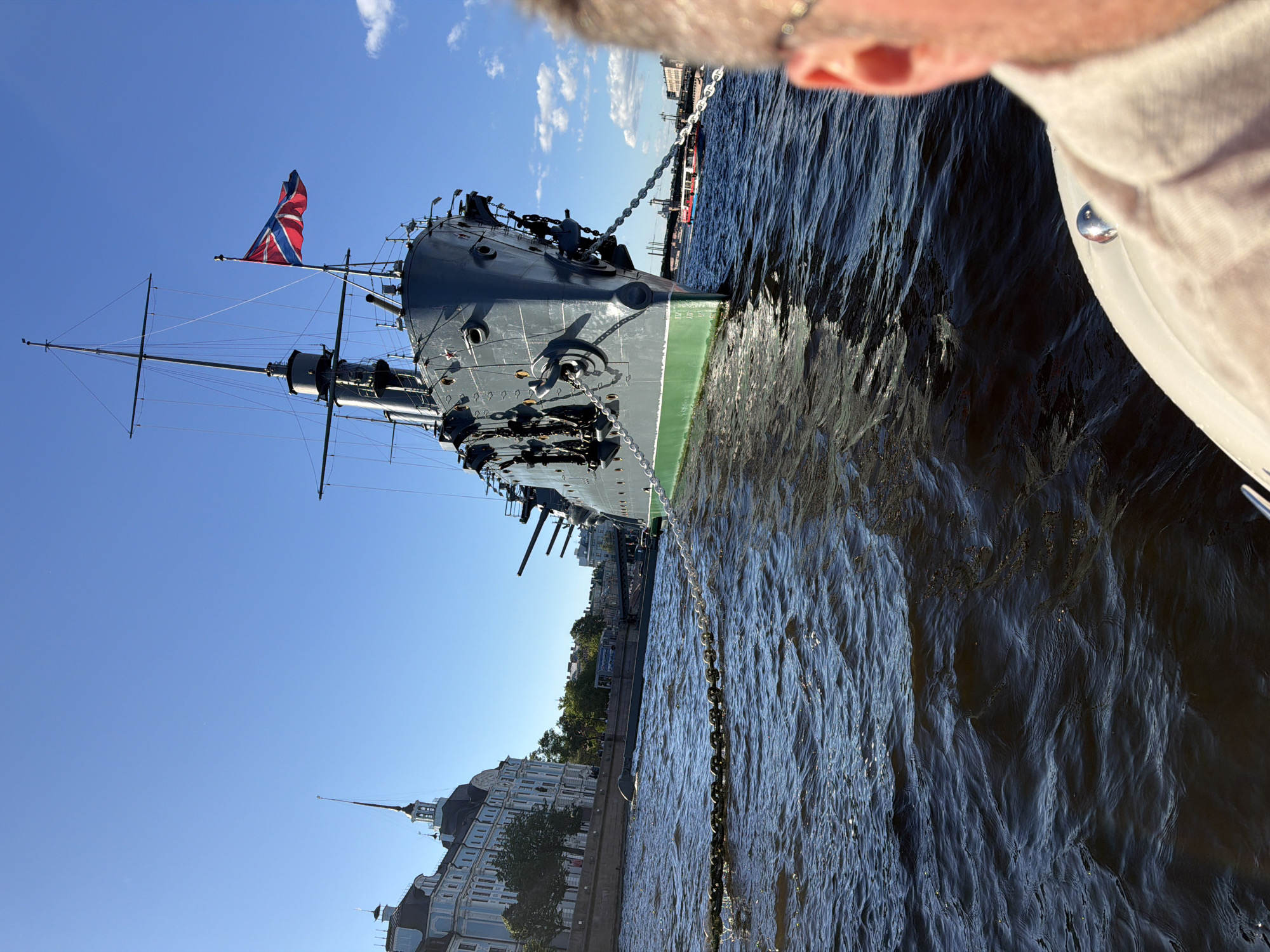 The image shows a view from a boat on the Neva River in Saint Petersburg, Russia. The primary subject is the historic cruiser Aurora, a museum ship and a significant landmark in the city. The Aurora is moored on the river, its grey hull and prominent gun turrets clearly visible under a bright blue sky. A Russian flag flies from its mast. To the left of the ship, the St. Petersburg State University of Economics and Finance, with its distinctive spire, can be seen along the embankment. The river is choppy, reflecting the sunlight. In the foreground, the back of a person's head and shoulder is visible, indicating the perspective of someone on another vessel. Several other boats and bridges are visible in the background, suggesting a busy waterway.