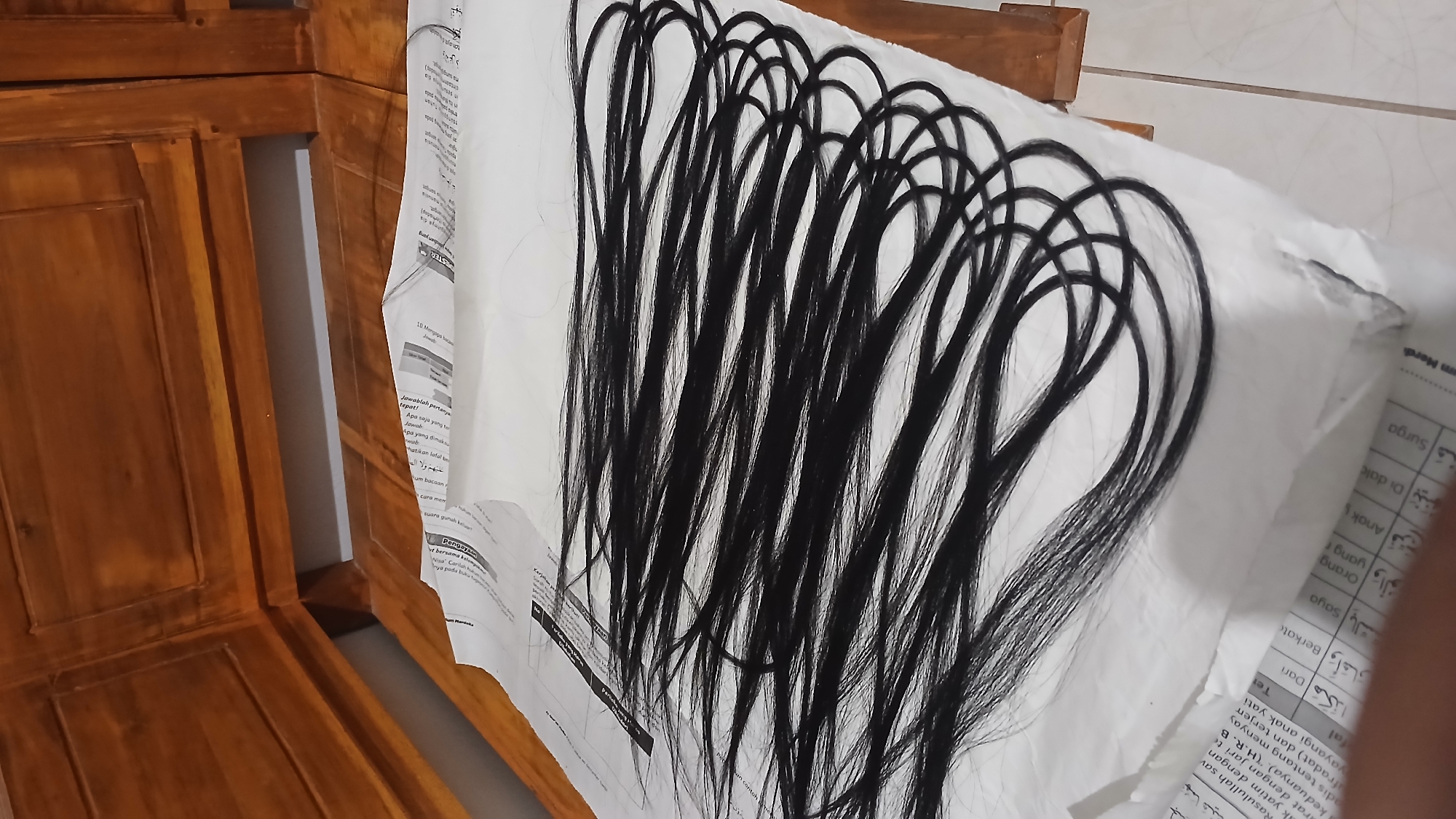 A close-up, low-angle shot shows several strands of long, black hair laid across a piece of white paper, which is itself resting on a wooden surface. The wooden surface appears to be part of a piece of furniture, possibly a table or bed frame, with visible grain and a warm, reddish-brown hue. The hair is dark and appears to be synthetic or human hair extensions, laid out in parallel lines, with some strands curled into loops. The paper underneath has text and some Arabic script visible, suggesting it might be a page from a book or newspaper. The overall lighting is dim, casting soft shadows and creating a muted atmosphere. The background is blurred, but hints of more wooden furniture suggest an indoor domestic setting. The scene does not contain any people or animals, nor does it depict any specific action beyond the arrangement of the hair. The location in Sampang, Indonesia, is not directly evident from the visual cues in the image.