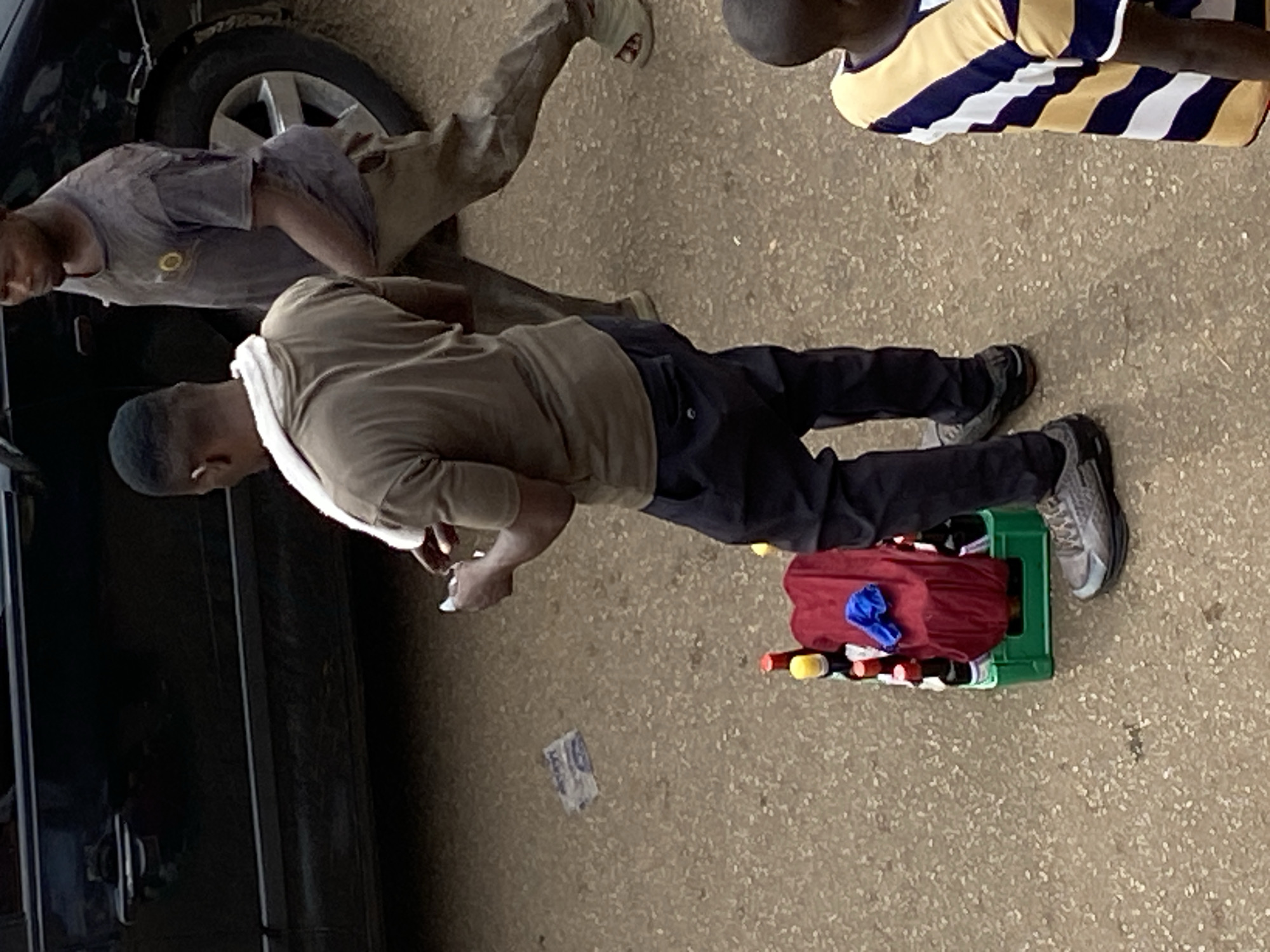 The scene appears to be on a street in Bukuru, Nigeria. A man stands next to a parked black vehicle. He wears a brown shirt, dark pants, and grey sneakers. A white cloth is draped around his neck. He holds something in his hands and looks down. Next to him on the ground is a green plastic crate containing bottles, mostly covered by a red bag with a blue object on top. Another person wearing a grey shirt and khaki pants is partly visible near the car's tire. A third person is seen in the upper right corner wearing a striped shirt. A single paper bill lies on the ground near the crate. The environment is a paved surface with a gritty texture.