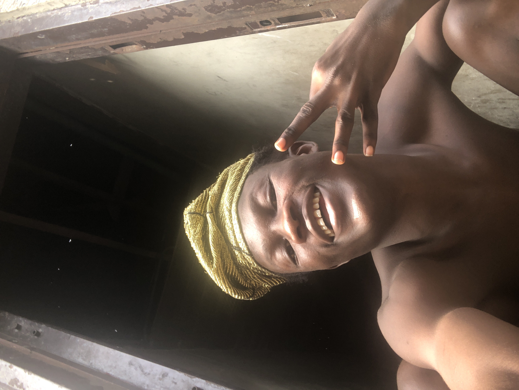 The media file shows a close-up, low-angle shot of a person with dark skin. The person is wearing a gold, beaded headscarf and has their eyes closed, with a wide smile revealing their teeth. One hand, with fingers extended in a peace sign gesture, is placed on the person's forehead near their eye. The background is dark and out of focus, with some lighter, textured surfaces visible at the top and bottom, possibly suggesting an indoor or sheltered outdoor space. The lighting appears to be natural, casting highlights on the person's skin and suggesting it might be daytime. The overall mood is cheerful and playful. No text is visible in the image. Given the context of Ado, Nigeria, the scene could depict a moment of personal expression or celebration in a local setting.