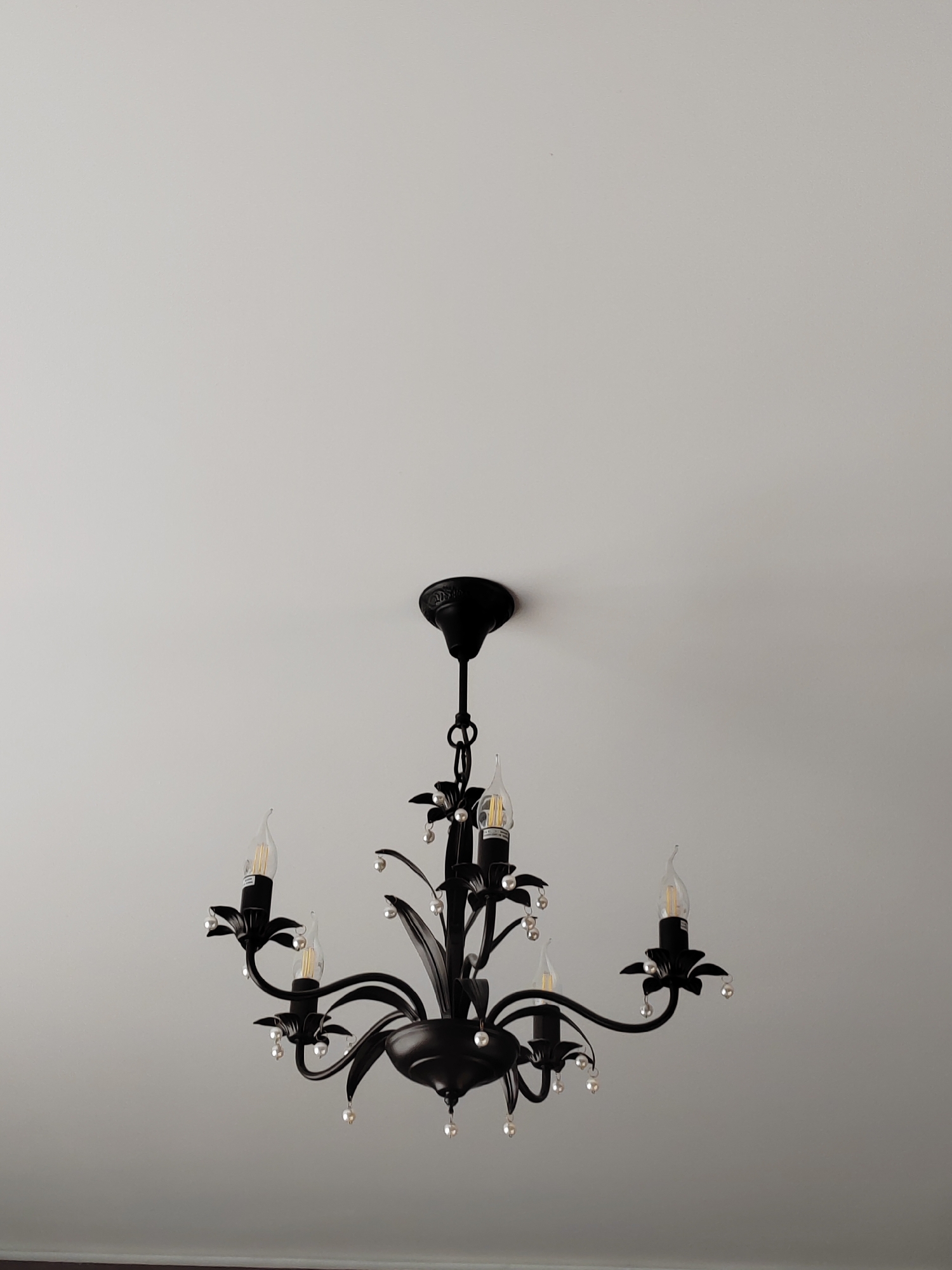 A black, five-light chandelier with faux-candle bulbs and clear crystal embellishments hangs from a white ceiling. The chandelier has ornate, curved arms and decorative floral elements at the base of each light. The ceiling appears to be a plain white surface, with no other visible features. The lighting in the image is soft and diffused, suggesting it may be daytime or the chandelier is not illuminated. The image is framed from a low angle, looking up at the chandelier. There are no people or other objects present in the scene. The location of the image is specified as Грачёвка, Russia.