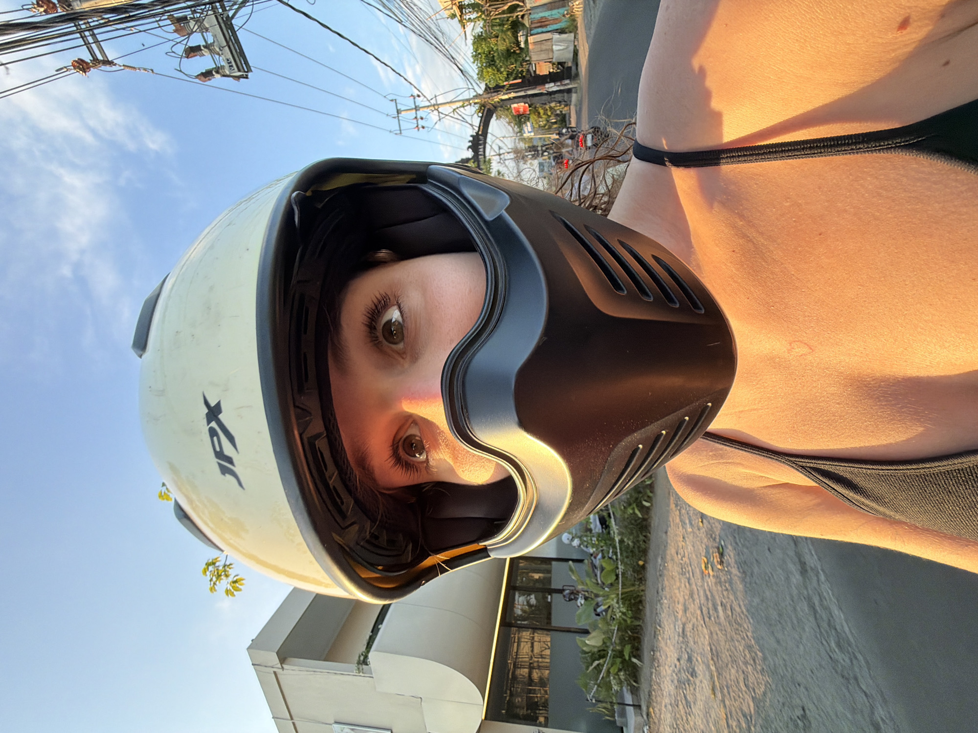 A close-up, low-angle, eye-level selfie shows a young woman wearing a tan and black full-face motorcycle helmet. She is on a street in North Kuta, Indonesia. The woman's eyes are visible through the helmet's visor. She is looking towards the camera. The woman is wearing a black bikini top. The sun is setting, casting a warm glow on her skin and the environment. The background shows a building with modern architecture, a street with power lines, and some vegetation. The woman appears to be riding a motorcycle or scooter, as suggested by the helmet and the street setting. The scene conveys a sense of travel and exploration in a tropical location.