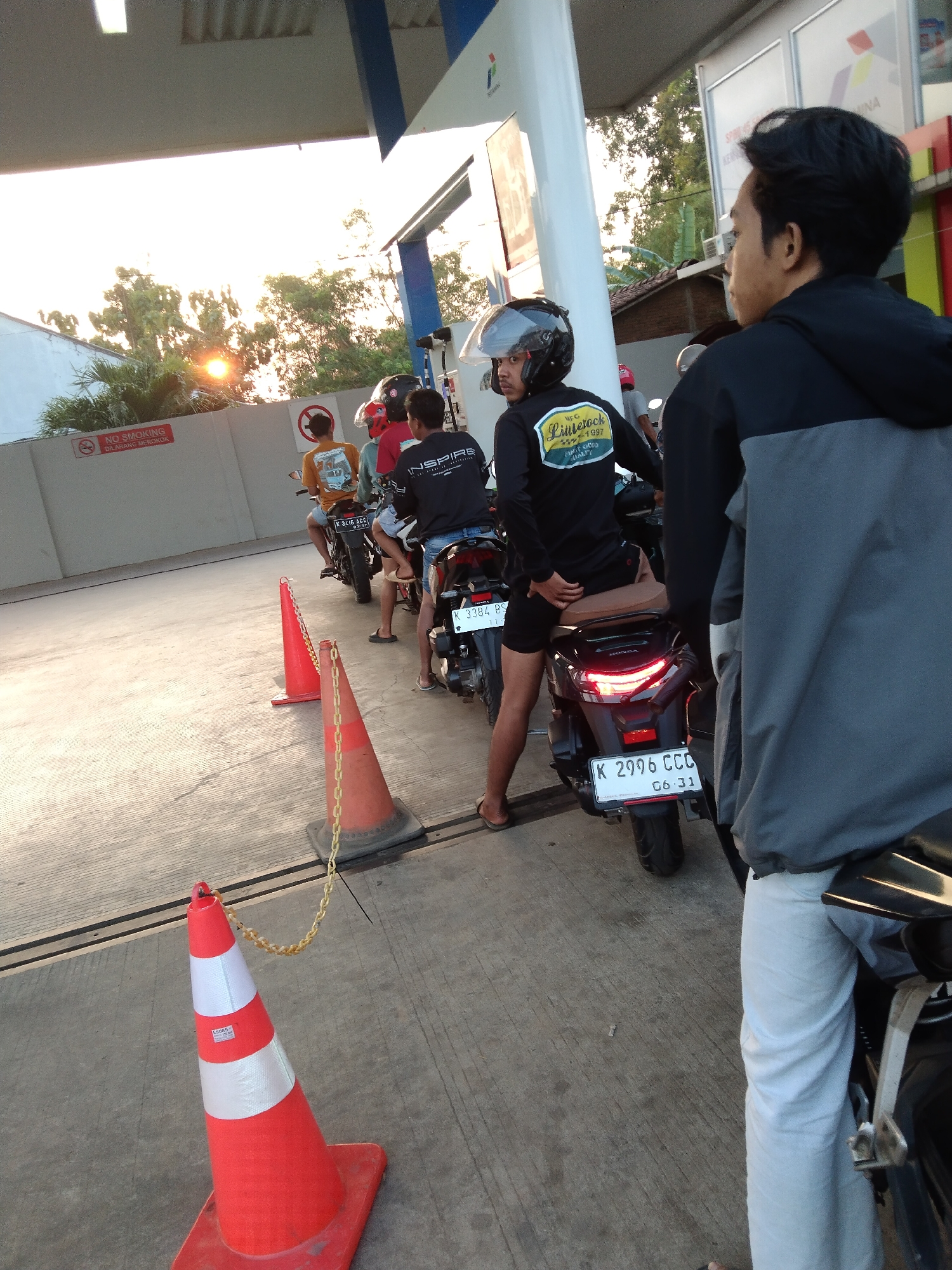 In Kembang, Indonesia, at a gas station, a line of motorcyclists waits. The foreground features an orange traffic cone with reflective stripes, its chain dangling. Behind it, a motorcycle with license plate K 2996 CCC and another with K 3384 BS are visible. The riders, all male, are dressed in casual attire. One rider in the foreground wears a black hoodie and light-colored pants. The background shows trees, a building, and the sun setting, casting a warm glow on the scene. A "NO SMOKING" sign is also present on a wall.