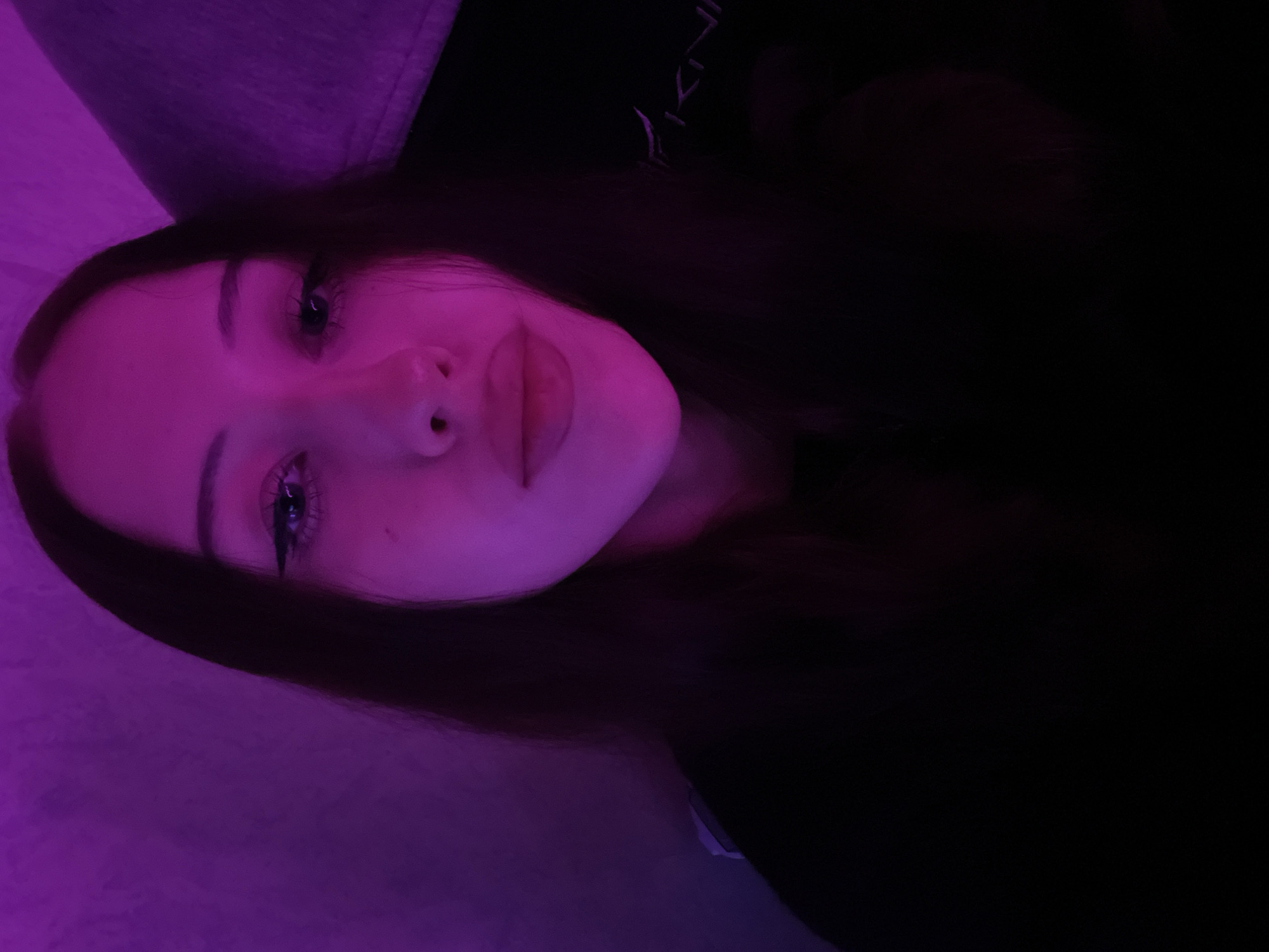 An image captured in the city of Votkinsk, Russia, displays a young female subject positioned centrally in a medium-close-up shot. Her long, dark, straight hair is parted on the viewer's right, framing her face and extending over her shoulders. The scene is dominated by a strong purple-magenta ambient light, primarily illuminating the left side of her face and the background, creating high contrast with the darker areas. She looks directly at the camera with dark eyes, defined by visible eyeliner and dark pupils. Her eyebrows are dark and arched, and her lips are full and closed. She is wearing a dark, long-sleeved garment, possibly black. The background on the viewer's left appears to be a textured wall, uniformly purple from the lighting. On the viewer's right, a darker, upholstered object, possibly a gaming chair or similar furniture, is partially visible, featuring faint, stylized text resembling "SKIN" or "IKIN" near the bottom right corner.
