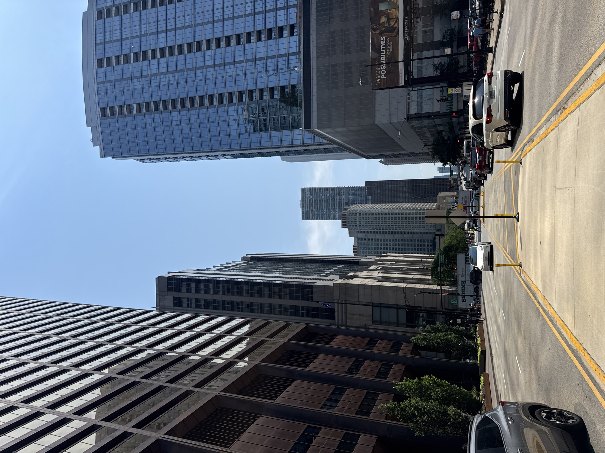 The image depicts a street view in Chicago, Illinois, United States of America. The scene is characterized by tall buildings on both sides of a wide, multi-lane road. The architecture suggests a modern urban environment with glass and steel skyscrapers dominating the skyline.
On the left side of the road, a dark-colored building with a grid-like facade, likely made of metal and glass, rises high into the sky. Adjacent to it, another building with a similar dark facade is visible. Further down the street, more buildings of varying heights and architectural styles can be seen.
On the right side, a sleek, modern skyscraper with a blue glass exterior is prominently featured. Below this skyscraper, a lower-rise building with large glass windows and a billboard is situated. The billboard displays an advertisement with the text "Pursue POSSIBILITIES" and an image of a person working at a desk.
The road itself is asphalt and appears to be a main thoroughfare with yellow double lines marking the lanes. Several vehicles are present, including cars, SUVs, and a truck. Some cars are in motion, while others appear to be stopped, possibly in traffic or at an intersection. Pedestrians are visible on the sidewalks, suggesting active street life. The sky is clear and blue, indicating a bright, sunny day. Green trees are planted along the sidewalks, adding a touch of nature to the urban landscape.
The perspective of the photograph is from street level, looking down the road, with the camera positioned between the sidewalk and the traffic lanes. The overall impression is one of a bustling, contemporary city.