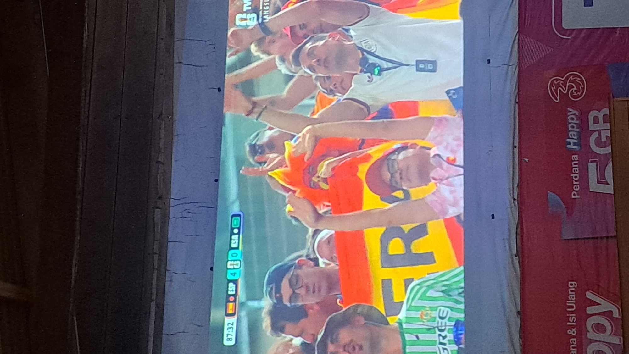 The image captures a live broadcast of a football match, displayed on a screen. The score visible on the screen indicates a match between ESP and KSA, with ESP leading 4-0 at 87:32. The scene on the screen shows a crowd of enthusiastic spectators, many waving flags and raising their hands in support. A large Spanish flag is prominently displayed, featuring the word "ESP" in large letters. The spectators are diverse, with various ages and styles of dress. The broadcast also shows a logo for "TV" in the upper right corner. Below the screen, a red banner is visible with text including "Perdana Happy" and "3 GB," suggesting it is a promotional banner from a telecommunications company in Payagaboh, Indonesia. The overall environment appears to be a public viewing area or a sports bar.