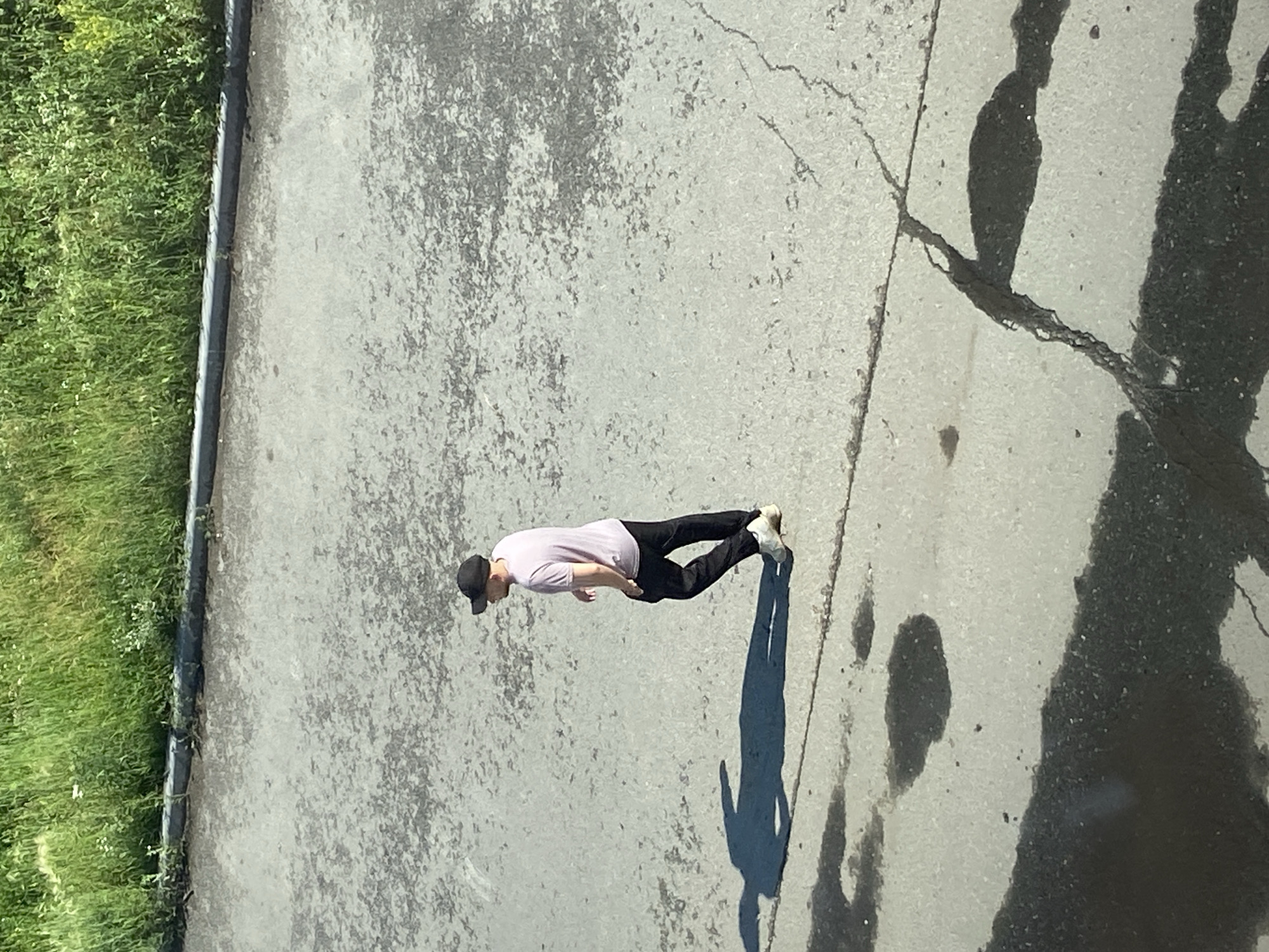 A man wearing a light pink t-shirt, black pants, and a black baseball cap walks across an asphalt surface near a patch of green grass. His shadow is cast to his left, elongated by the sun. The asphalt appears somewhat cracked and weathered, with several dark patches that could be oil stains or wet spots. The scene is set outdoors, with the grass suggesting an area that is not fully urbanized, potentially a park, verge, or undeveloped lot within the city of Zavodoukovsk, Russia.