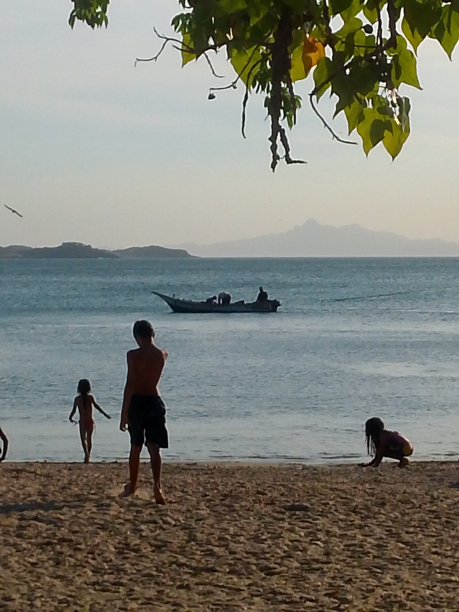 The image depicts a beach scene in Juan Griego, Venezuela, featuring four individuals: one child is walking in the water towards the left, a second child is in the foreground, a third is crouching on the sand to the right, and a fourth obscured to the left. A small boat with two people aboard is visible in the water, with a mountain range in the background. Tree branches and leaves are partially visible at the top of the frame.