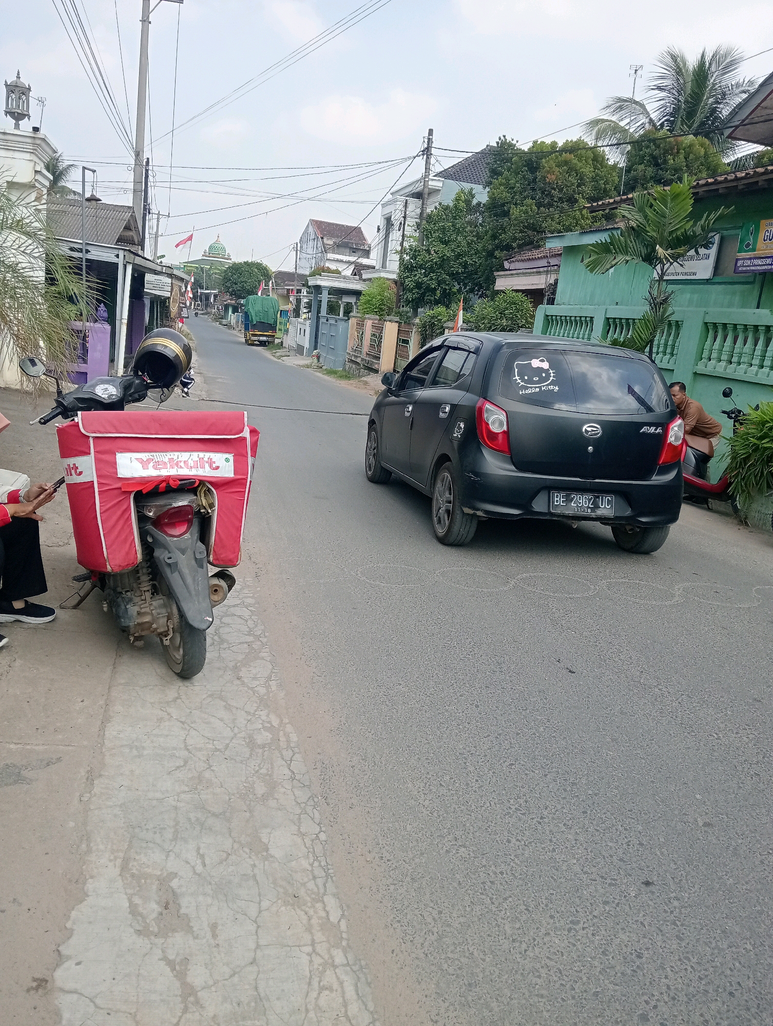 A black Daihatsu Ayla hatchback is parked on a street in Pringsewu, Indonesia. The car has a "Hello Kitty" sticker on the rear window. A red motorcycle with a large "Yakult" delivery box is parked next to the car. A person is sitting on the sidewalk next to the motorcycle, looking at their phone. The street is lined with buildings and trees, and there are power lines overhead. The sky is overcast.