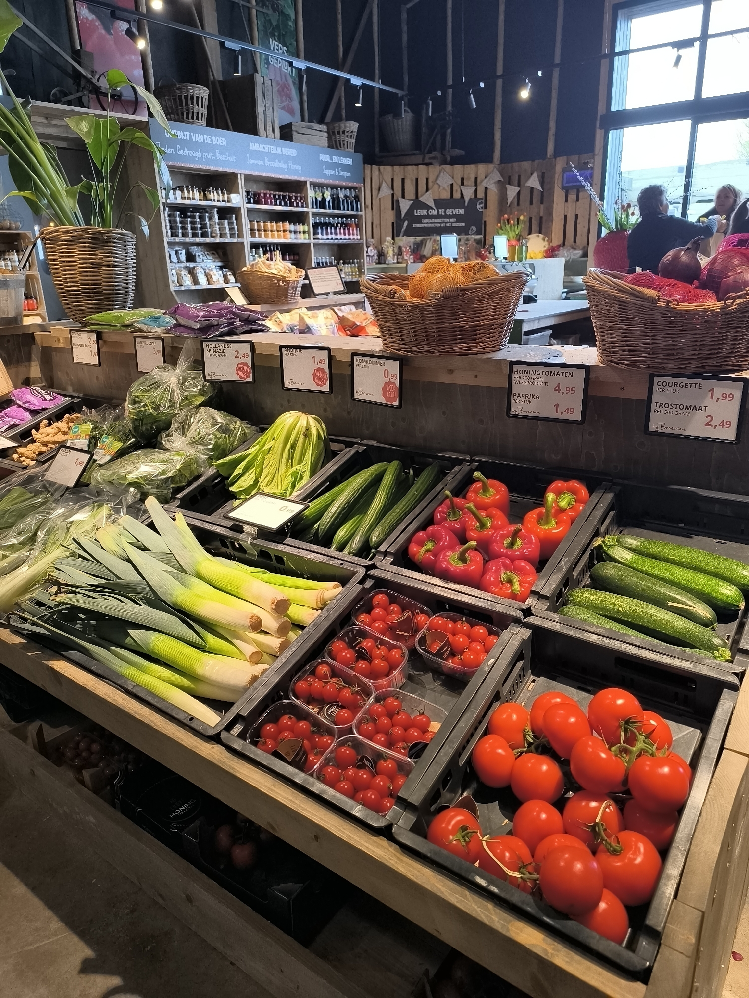Here is a detailed description of the image:
**Scene:** An indoor produce market in Warmenhuizen, Netherlands.
**Subjects/Objects:**
* A variety of fresh vegetables are prominently displayed in black plastic crates on a wooden counter. These include:
* Leeks
* Cucumbers
* Red bell peppers
* Zucchinis (Courgettes)
* Cherry tomatoes
* Tomatoes on the vine
* Other vegetables (spinach, broccoli and onions) are displayed above
* Price tags indicating the cost of the produce.
* Behind the produce display is a shelf unit containing bottled goods, jams and preserves.
* Baskets containing what looks like bread and onions.
* There is a large leafy plant in a woven basket to the left.
* In the background, there is a window with daylight coming through, as well as people.
**People:**
* There appear to be two women standing together in the background near the window.
**Actions:**
* The produce is arranged for sale.
* Customers are shopping (implied by their presence in the background).
**Environment:**
* The market has a rustic feel with exposed wooden beams and shelving.
* The lighting is a mixture of natural daylight and artificial lights.