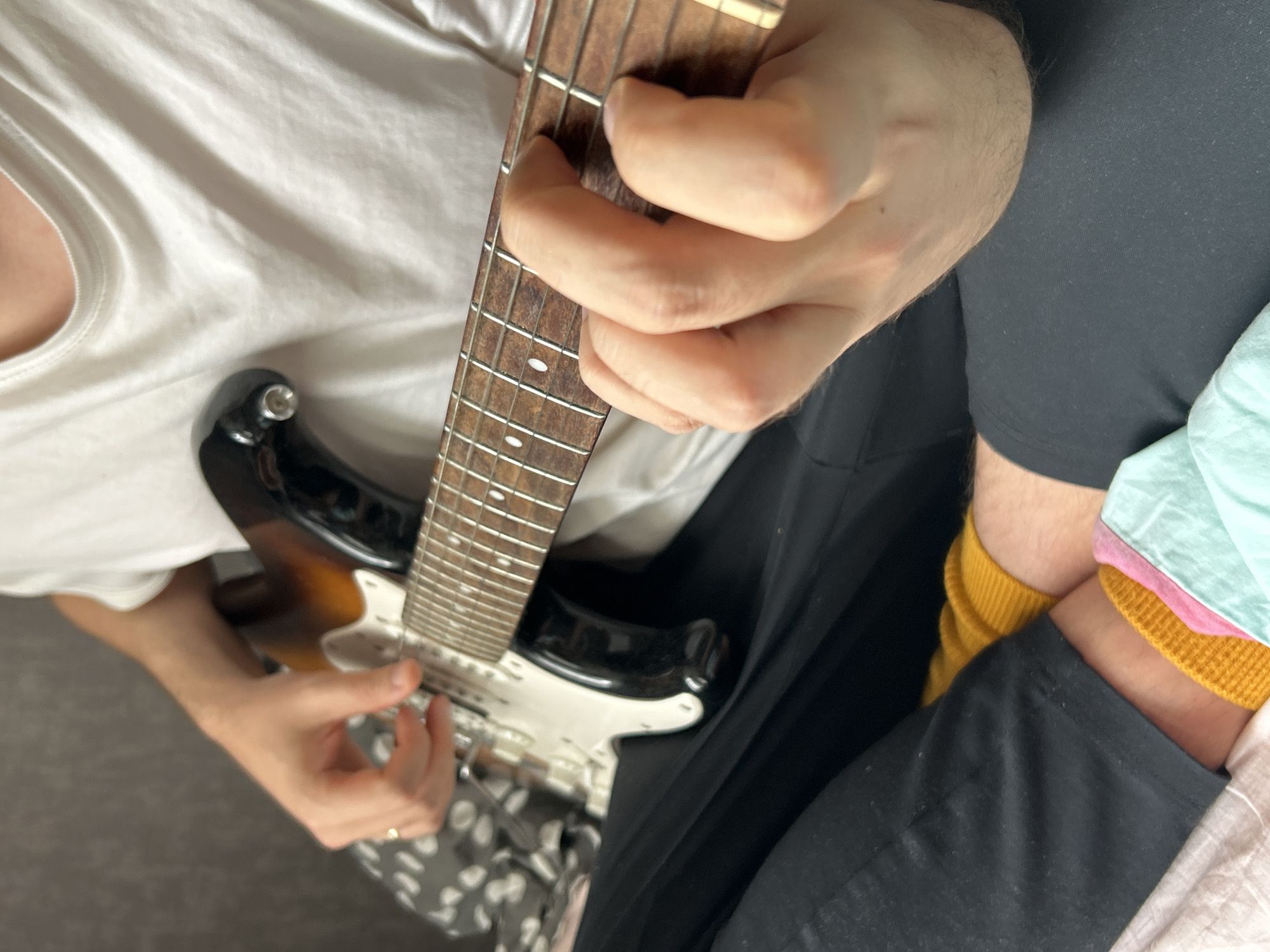 A person wearing a white t-shirt and black pants is holding and playing an electric guitar. Their left hand is fretting the strings near the guitar neck, while the right hand is positioned near the body of the guitar. The guitar has a sunburst finish. The person is also wearing yellow socks. The location is unknown.