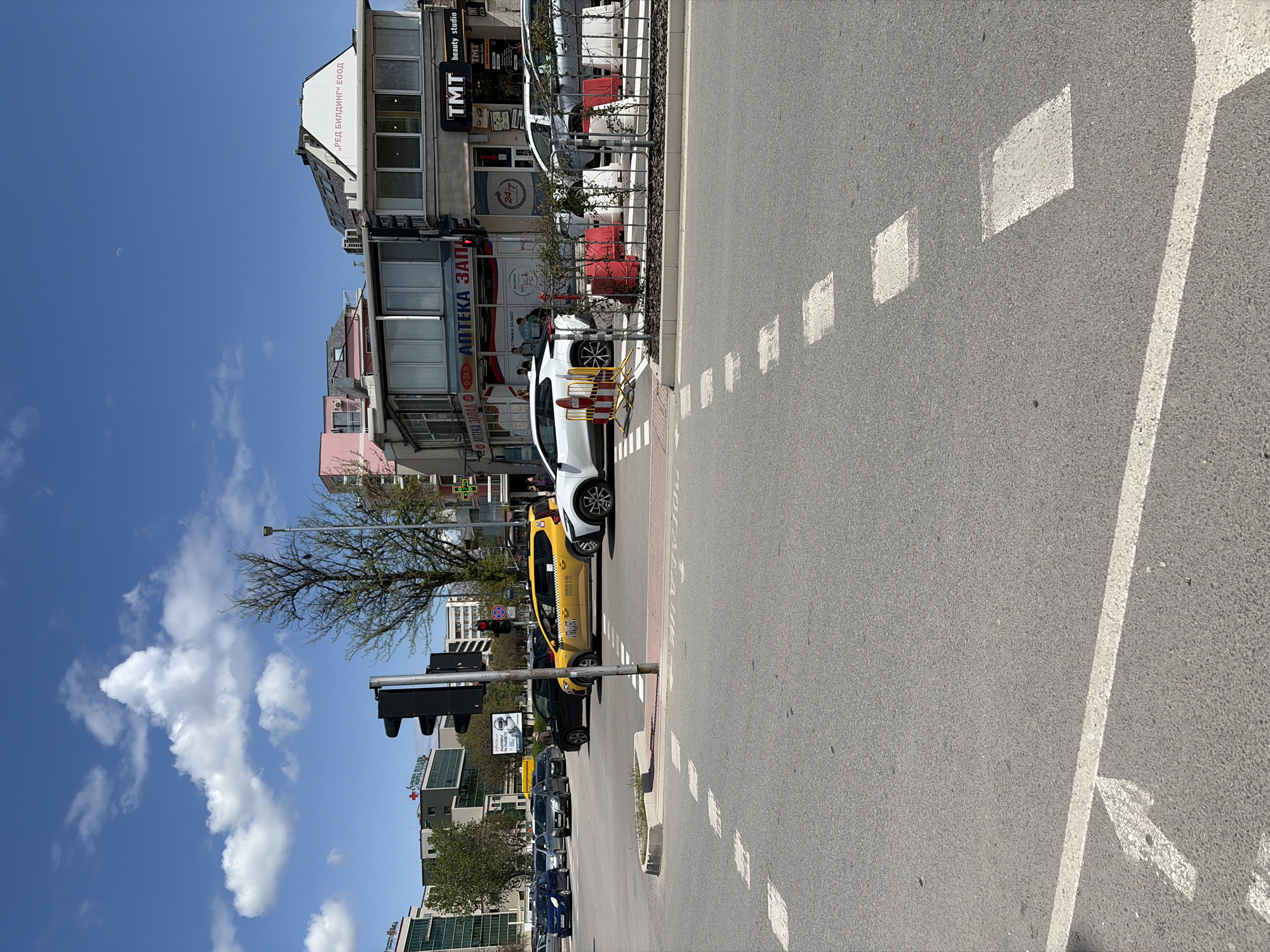 An urban street scene unfolds on a clear, sunny day in Plovdiv, Bulgaria. The original image is rotated counter-clockwise. The asphalt road occupies the foreground and midground, featuring distinct white road markings including dashed lines indicating lanes or pedestrian crossing zones, a solid white line, and a white arrow painted on the surface.
To the left of the road, a sidewalk is lined with parked vehicles. A prominent yellow taxi, branded "ТАКСИ" with a phone number "0888 88 11 90" on its side, is stopped or parked. Ahead of it, a white car, possibly an SUV, is also parked near a pedestrian crossing marked with red and white bars and yellow traffic cones/barriers. Further down the street, several other cars, including a black and blue one, are visible. Traffic lights and a lamppost stand along the street, and a tree with sparse foliage is present.
The background is dominated by multi-story buildings housing various businesses. Visible text on storefronts includes "АПТЕКА ЗАЛАЛД" (a pharmacy, one sign also displays "24/7"), "РИЖА ЗА ВАС!", and "TMT beauty studio" and "TMT" on dark signs. Another building prominently displays "„РЕД БИЛДИНГ" ЕООД". A red cross symbol is also visible on a building further in the background, possibly indicating another medical facility.
The sky is bright blue with scattered white cumulus clouds. A faint crescent moon is discernible in the upper left portion of the sky, suggesting it's daytime, likely mid-morning or afternoon. The overall atmosphere is that of a bustling, functional urban environment.