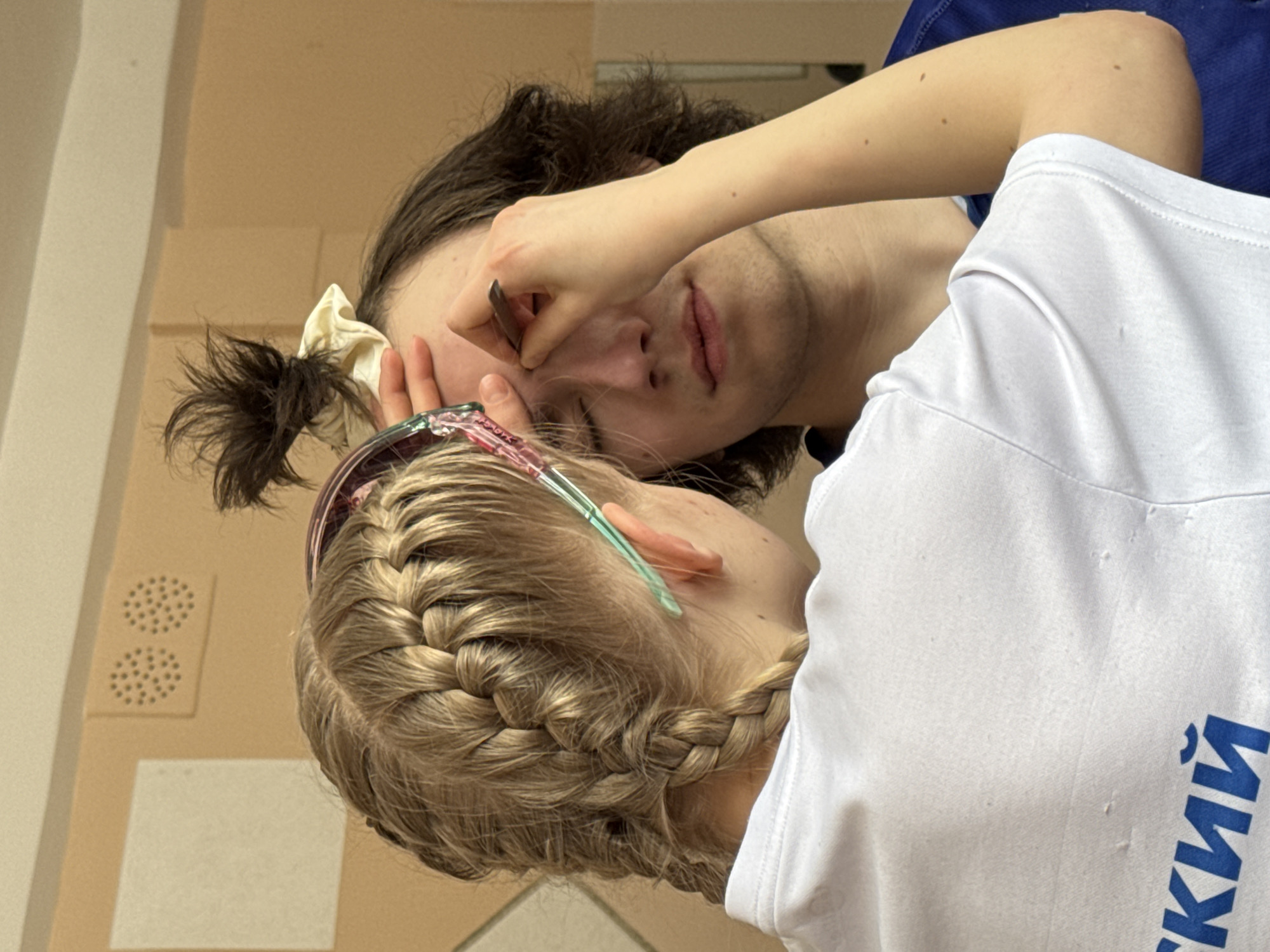 A young woman with blonde braids is using tweezers to pluck the eyebrows of a young man with dark hair, who is lying down. The environment suggests an indoor space, possibly a home, given the peach-colored walls and a white panel on the left. The location is unspecified, and there's no information in the visual details to confirm its connection to Berezniki, Russia.