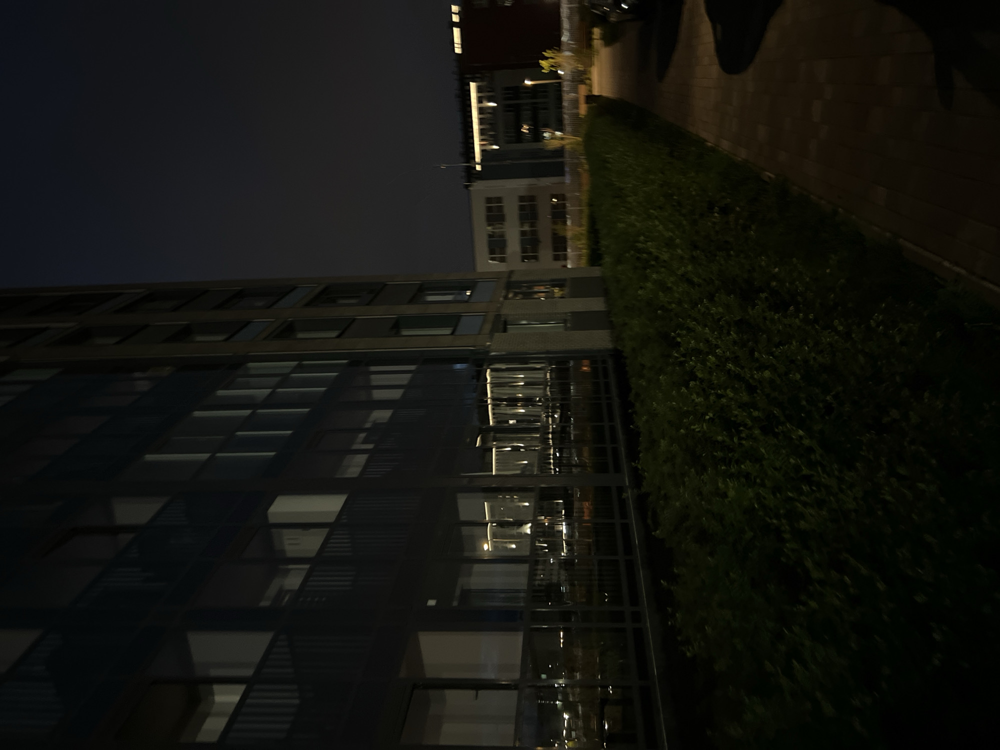 An exterior night shot shows a modern apartment building with large windows reflecting lights. In the foreground, a low, dense green hedge runs along the base of the building. To the right, a paved walkway with a brick pattern recedes into the distance. Further back, other buildings are visible, with lights on in some windows and illumination from streetlights or exterior building lights. The sky is dark, indicating nighttime.