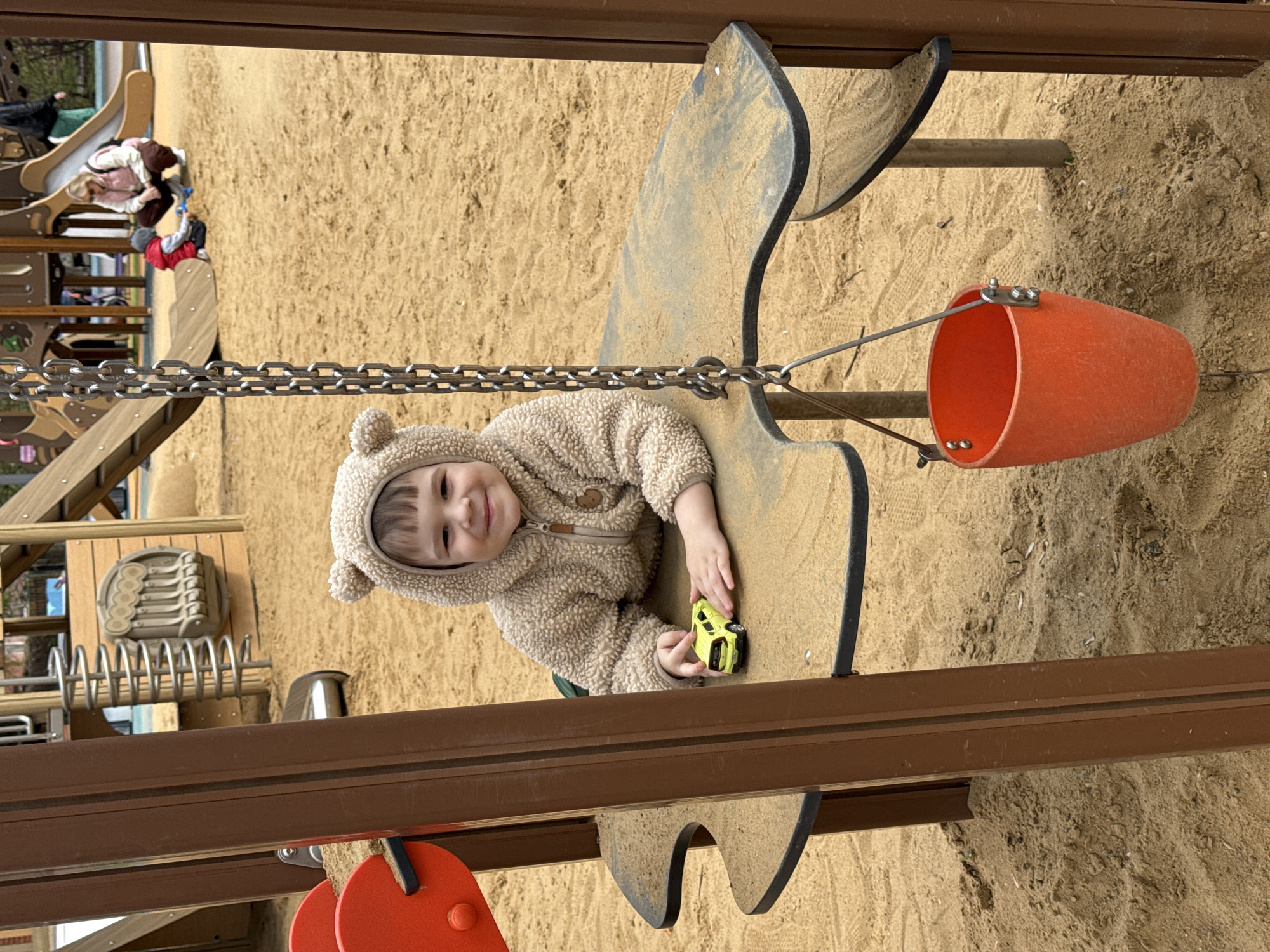 A child in a light-brown, teddy-bear-style hooded jacket sits in a piece of playground equipment, holding a small yellow toy car. The playground appears to be sand-covered. An orange bucket hangs nearby. Other playground structures and people are visible in the background. The location is Moscow, Russia.