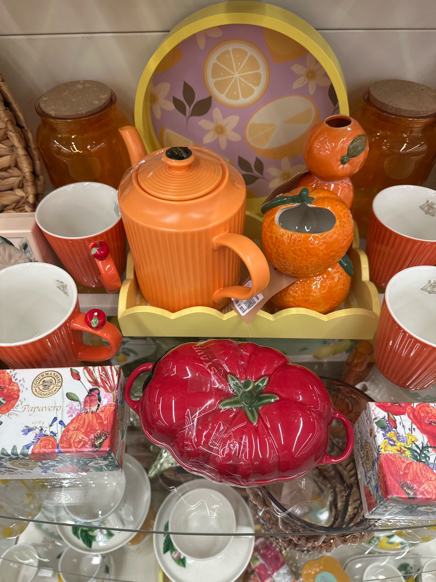 An eye-level, medium shot captures a display of decorative kitchenware on glass shelves. The items are primarily orange and red, with accents of white and yellow.
In the center of the frame, an orange teapot with a ribbed texture sits on a yellow scalloped tray. To the right of the teapot are two decorative orange pieces shaped like tangerines, one with a green leaf and stem, the other as a planter or sugar bowl. To the left of the teapot is a large, clear amber glass jar with a cork lid, and next to it, a woven basket handle peeks into the frame.
Arranged around the central display are several ribbed orange mugs with white interiors, each adorned with a small red apple on its handle.
In the foreground, a bright red ceramic tomato-shaped dish, wrapped in plastic, dominates the bottom half of the image. To the left and right of the tomato dish are square boxes with floral and bird patterns, featuring red poppies and yellow flowers.
Below the main display, glimpses of white ceramic cups and saucers with botanical designs are visible on lower shelves, along with more decorative boxes and what appears to be a brown woven tray.
The lighting is bright and even, highlighting the glossy finishes of the ceramic and glass items. The background consists of a plain, light-colored surface, likely a shelf or wall. The scene appears to be a retail display in a store, possibly a home goods or kitchenware shop.