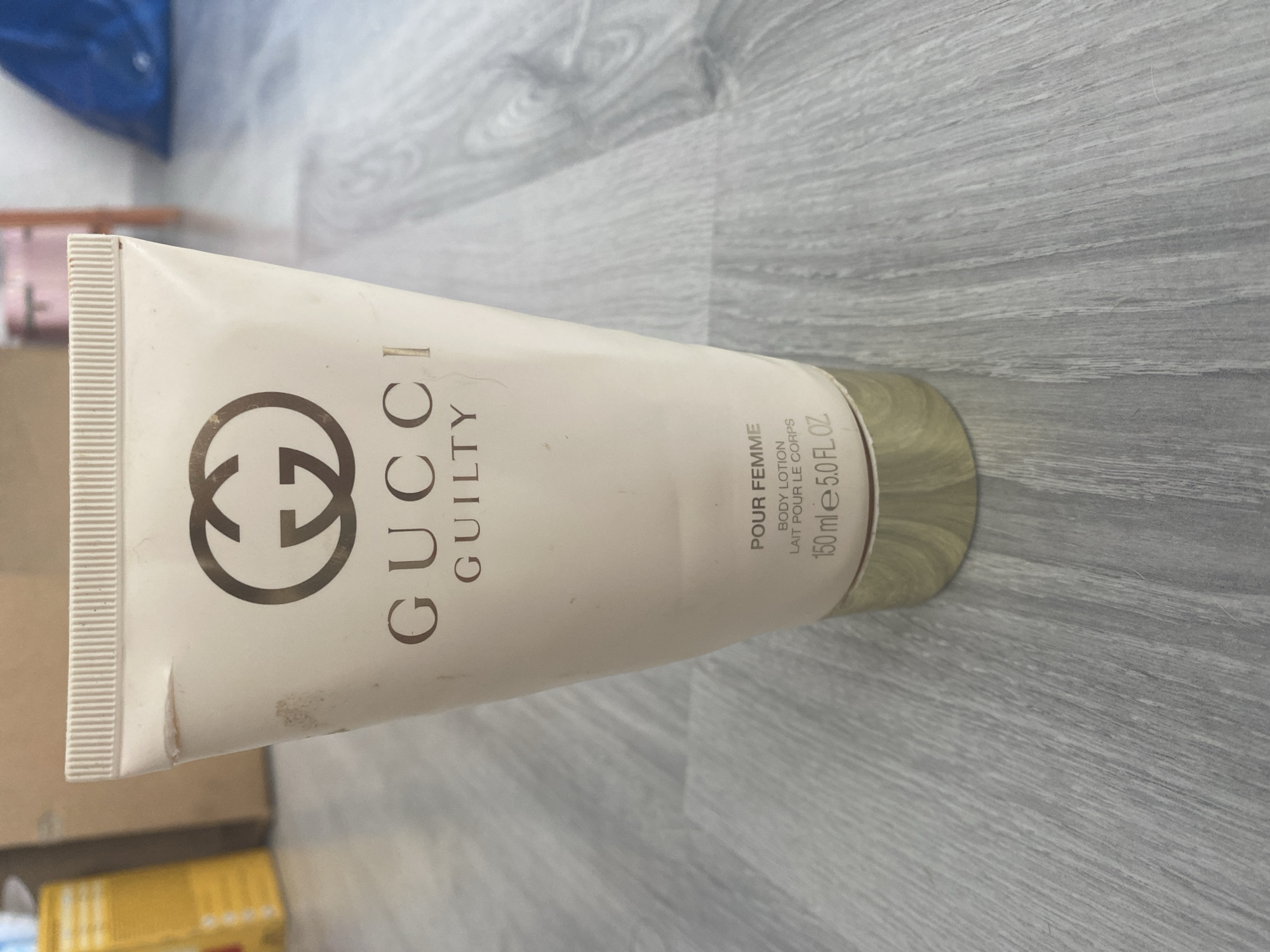 A close-up, slightly angled shot shows a pale pink tube of Gucci Guilty body lotion standing upright on a light gray wooden floor. The tube has a ribbed cap and a gold-colored base. The Gucci logo, a stylized "G" in gold, is prominently displayed in the upper half of the tube, above the text "GUCCI GUILTY." Below this, the text "POUR FEMME," "BODY LOTION," "LAIT POUR LE CORPS," and "150 ml e 5.0 FL.OZ." is visible. The tube has some light scuff marks and a small tear near the top. The background is blurred, showing what appears to be cardboard boxes and other indistinct objects, suggesting an indoor setting. The image was taken in Blagoevgrad, Bulgaria.