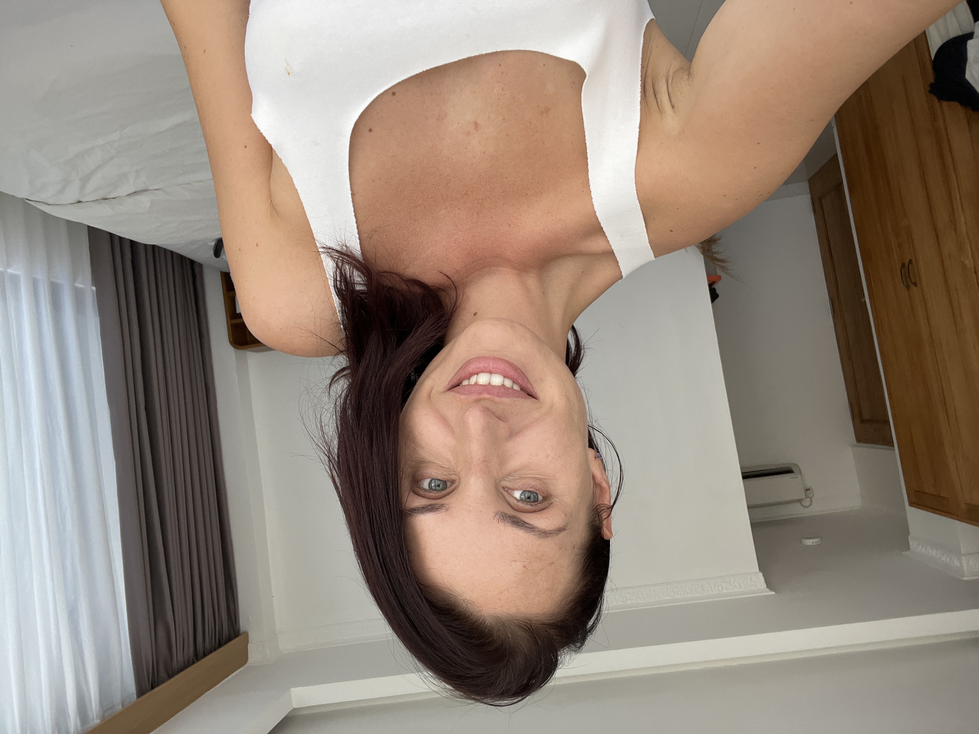 The image displays a woman, oriented upside down, taking a selfie and smiling directly at the camera. She has dark brown hair, which is styled to fall mostly to her left side, and light blue-green eyes. Her complexion is fair to lightly tanned, with noticeable freckles on her chest and face. She is wearing a plain white tank top or camisole.
The setting is an indoor room, likely a bedroom or hotel room, as indicated by the presence of a bed with white sheets on the left side of the frame. Behind the woman, to her left, are windows covered with white sheer curtains and darker grey-brown opaque curtains. To her right, part of a wooden wardrobe or cabinet with two doors is visible, alongside a white wall-mounted air conditioning unit. The room features white walls and light-colored flooring, with white crown molding adorning the upper wall section. A small, white, circular object lies on the floor near the air conditioner.
The activity taking place is a self-portrait, with the woman holding the camera above her head, looking downwards, and exhibiting a happy expression. The lighting suggests it is daytime, with natural light entering the room. The overall context places the scene in Kuta Selatan, Indonesia. No discernible text is clearly visible in the image.