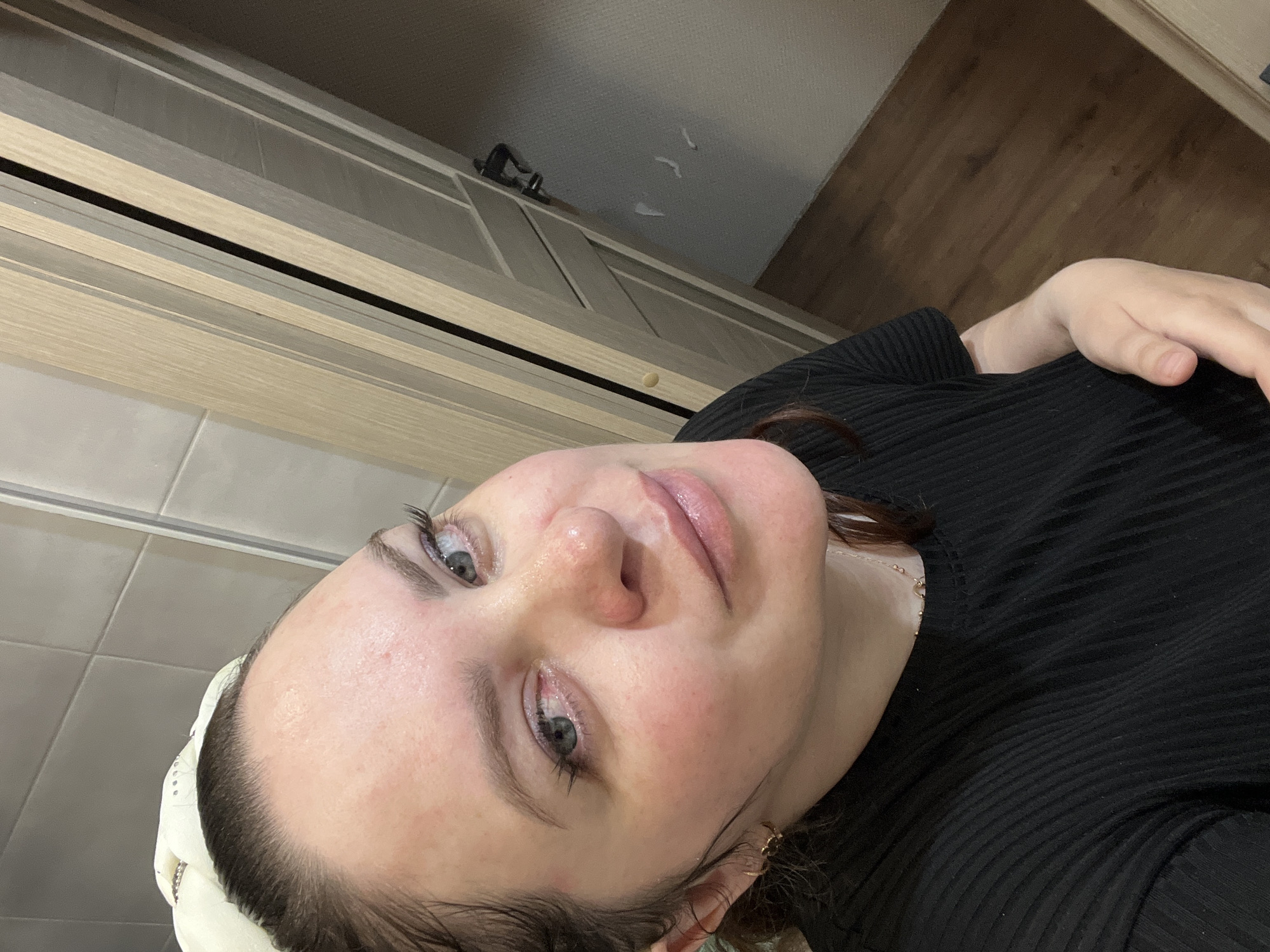 The image displays a female individual in a domestic interior located in Noviy, Russia. The subject is captured in a medium close-up, directly facing the camera with a subtle expression. Her skin is fair, exhibiting visible pores and localized erythema on the forehead and cheeks. Her dark hair is pulled back, partially covered by a light-colored head wrap or band adorned with circular embellishments. Her eyes are light blue-grey, featuring distinct eyelashes. Her lips are glossy with a natural pink hue. She wears a black, vertically ribbed short-sleeved top, a delicate gold-toned necklace with a small pendant, and a small gold-toned, possibly bow-shaped, hoop earring in her left ear. The background on the upper left consists of light grey tiled walls. To the right, a light-toned wooden door, characterized by vertical paneling, is partially open, revealing an adjacent interior space. This revealed space features an off-white wall with three small, irregular white adhered objects and a dark wooden floor. A dark fixture, possibly a door handle or wall hook, is visible on the far wall through the doorway. The subject's left hand is visible at the lower right, resting against her midsection.