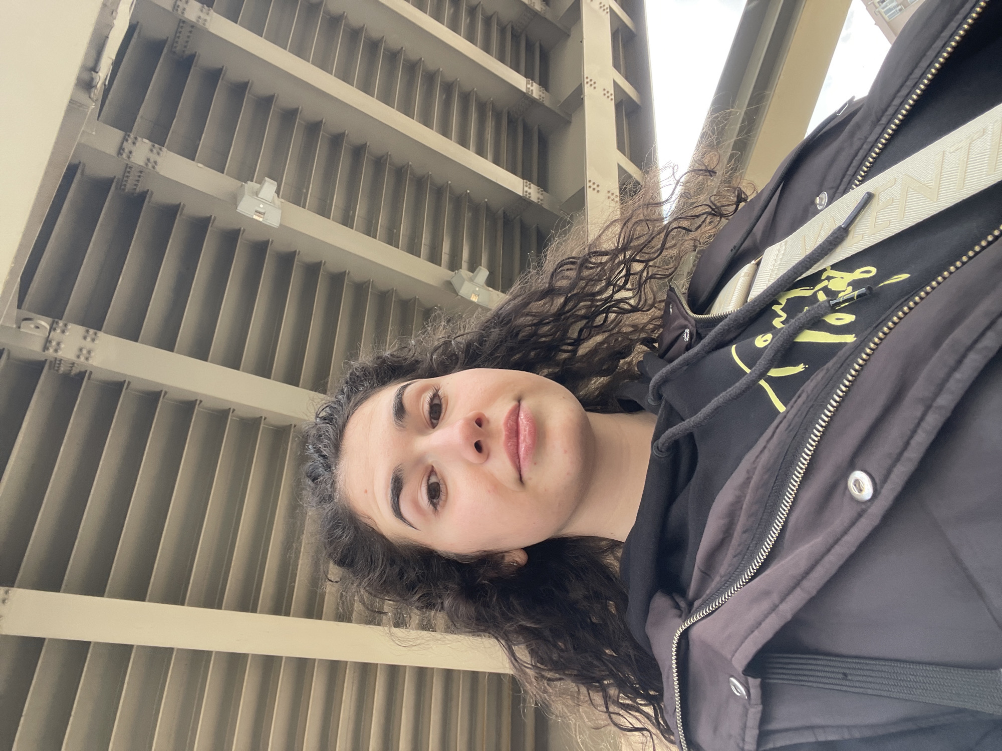 A young woman with curly brown hair is centered in the image, facing the camera with a slight smile. She is wearing a black hoodie with a yellow design underneath an open black jacket and a cream-colored strap across her chest. The background features a large structure with repetitive parallel beams, likely a bridge or overpass. The overall perspective suggests the photo was taken from a low angle, looking upwards. While the precise location within Sofia, Bulgaria cannot be determined without further contextual clues, the architectural style of the structure in the background, with its linear, repetitive design, could be indicative of an urban area in Sofia.