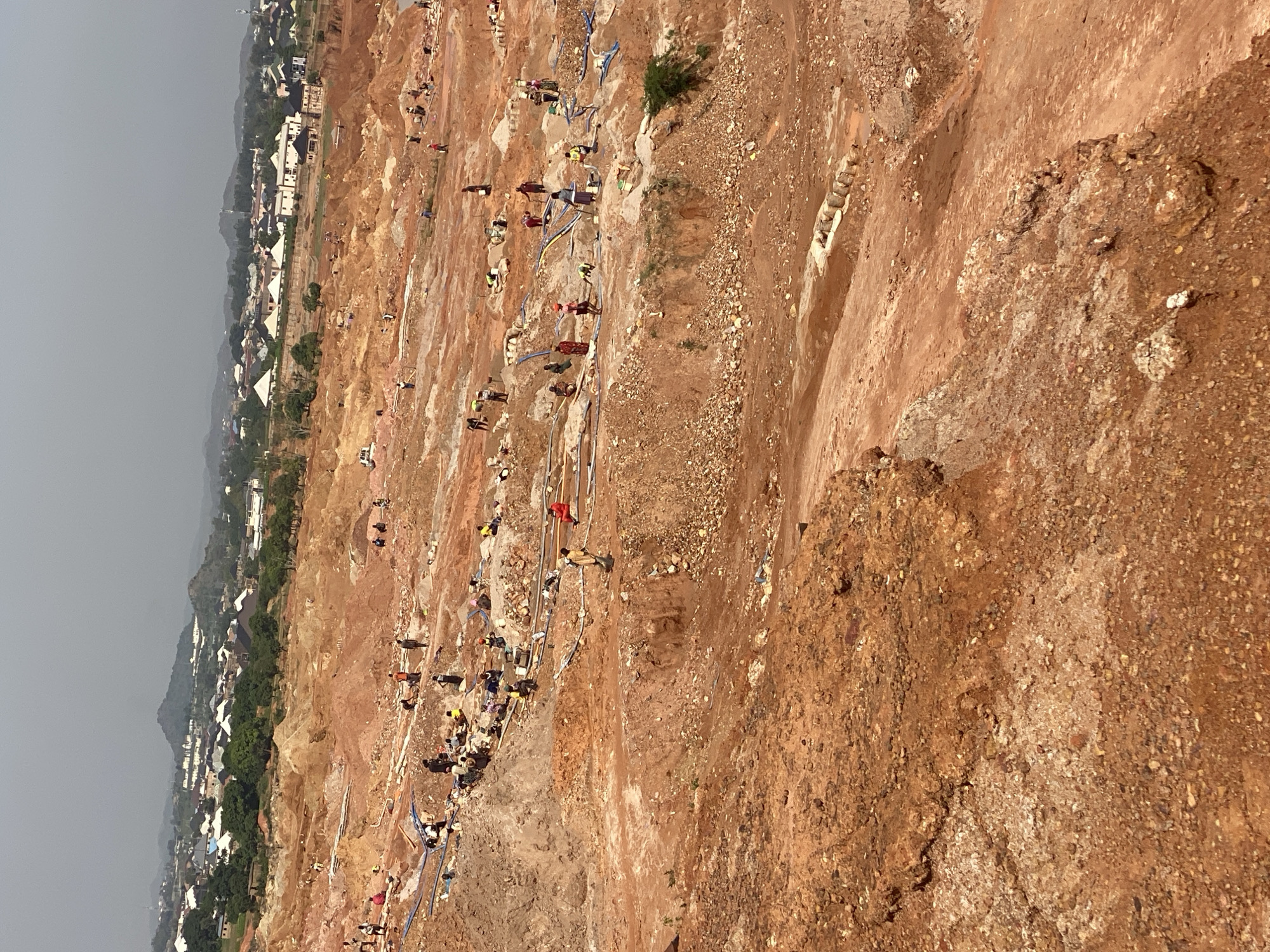 The image depicts an open-pit mining operation in Jos, Nigeria, characterized by a vast expanse of red earth, with numerous individuals engaged in manual labor. The terrain is uneven and excavated, revealing layers of soil and rock. A settlement, featuring buildings and vegetation, is visible in the background, suggesting the proximity of a populated area to the mining site. The people are dispersed across the site, some working individually and others in small groups. Their activities appear to involve digging, carrying materials, and managing water hoses, indicative of a labor-intensive extraction process. The overall environment is dusty and appears to be under a hazy sky.