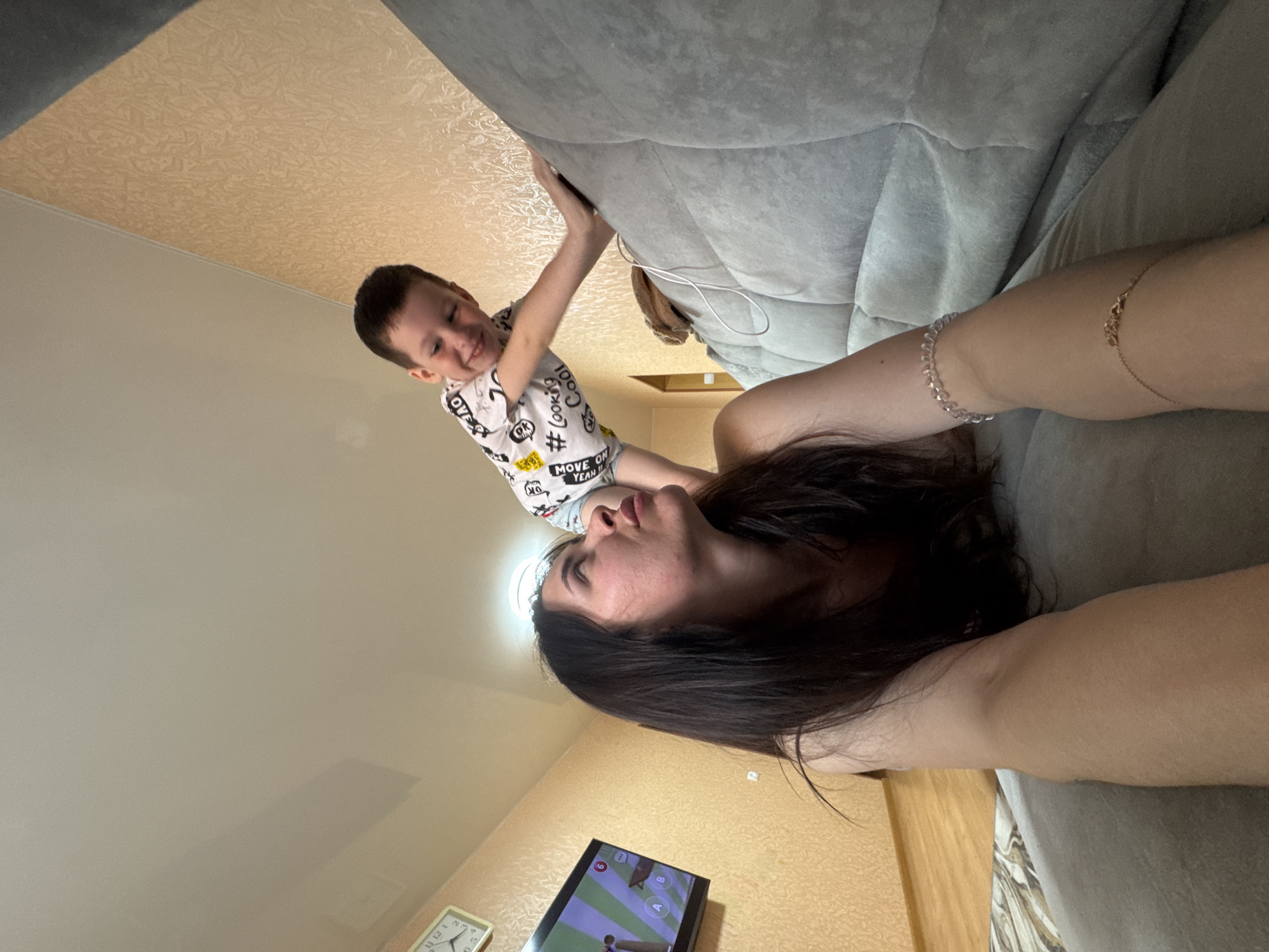 The image is a low-angle selfie taken indoors, showing a woman lying on a gray sofa. A young boy is positioned above her, appearing to lean over the sofa. The woman has dark hair and is looking towards the boy. She is wearing bracelets on her right wrist.
The boy is wearing a white t-shirt with black and yellow graphics and gray shorts. He seems to be holding onto the back of the sofa.
The background includes a patterned ceiling, a light-colored wall, and a television screen and a clock.
The image was taken in an apartment. The presence of Russian writing on the boy's t-shirt and in the TV screen indicate this may be located in Russia. The city where the picture was taken is Chaikovsky.