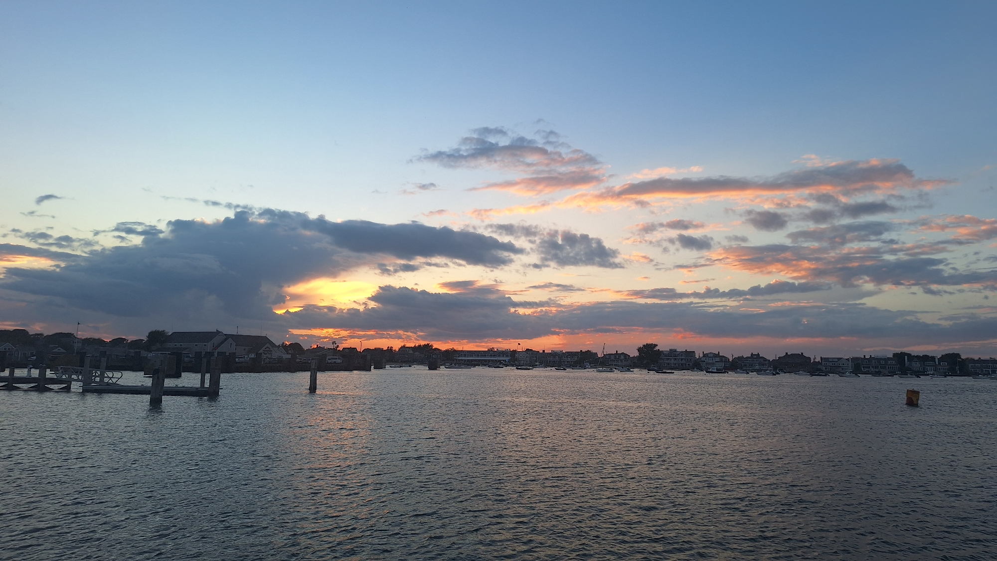 The image depicts a serene harbor scene on Nantucket, Massachusetts, USA, at dusk. The sky is a canvas of soft blues and pinks, with scattered clouds tinged by the setting sun. In the foreground, the calm, dark water of the harbor reflects the twilight sky. A wooden dock with several pilings extends into the water on the left side of the frame. Across the harbor, a picturesque waterfront is lined with a collection of houses and buildings, suggesting a coastal town. Numerous boats are anchored or moored in the harbor, adding to the maritime atmosphere. A yellow buoy is visible in the water on the right side. The overall impression is one of tranquility and the peaceful transition from day to night.